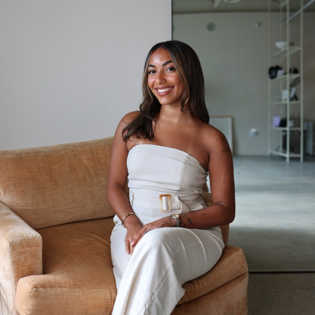 Woman in white strapless dress sitting on a beige sofa, smiling, with an indoor backdrop featuring shelves in the background.