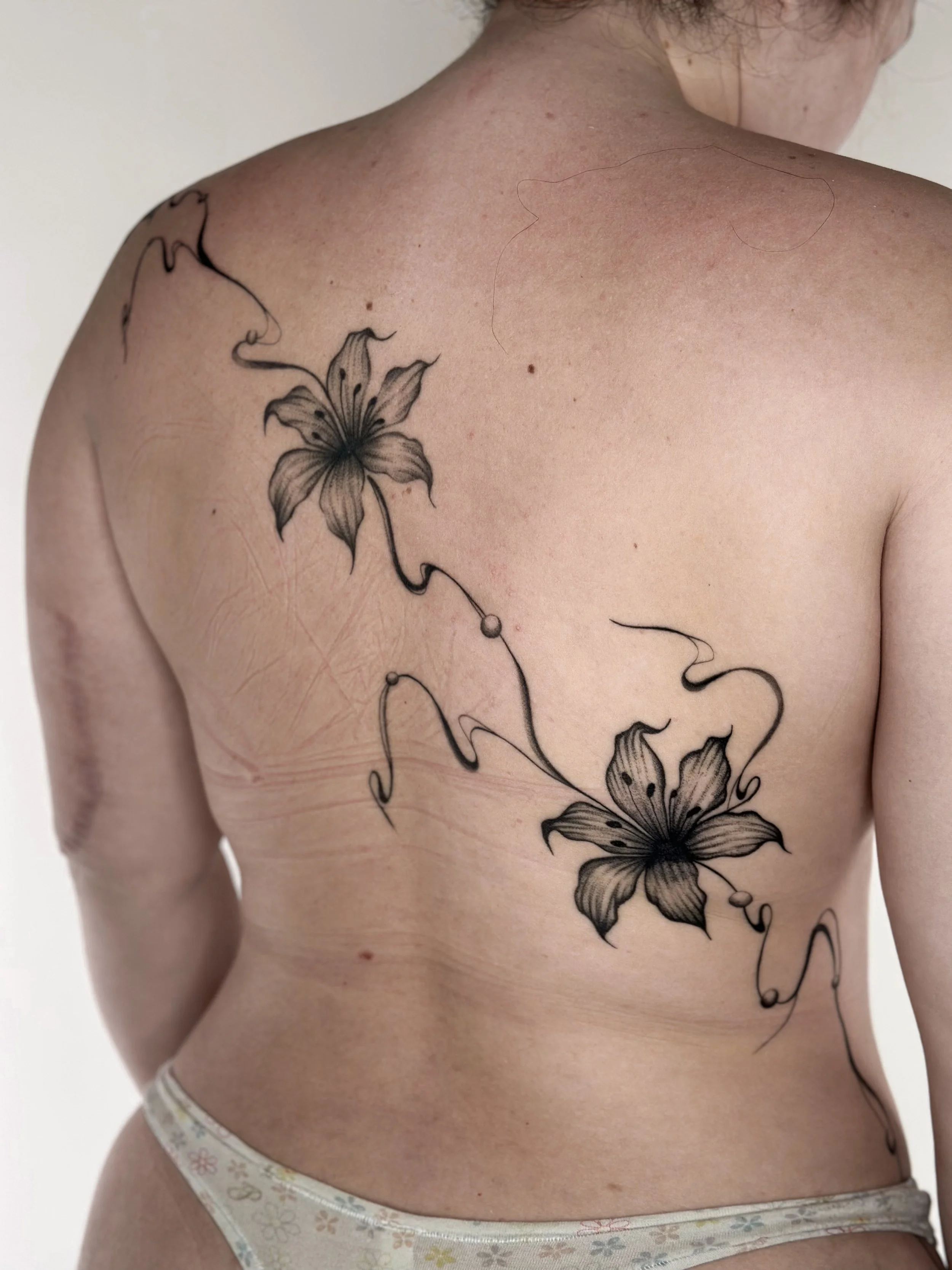 Black and gray floral tattoo of two lilies on a person's back, with flowing, curling stems and leaves.