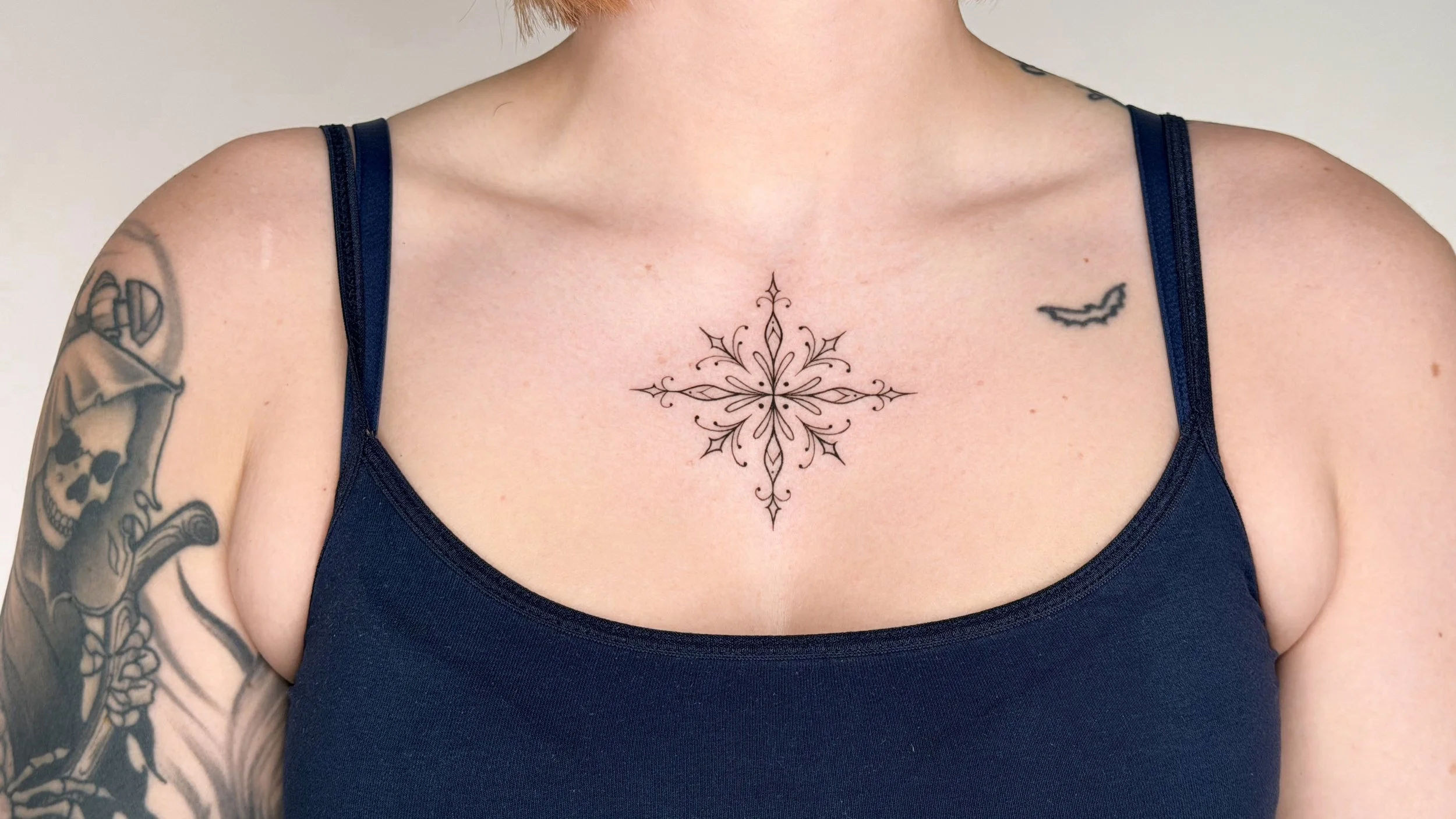 A woman wearing a navy blue tank top, showing tattoos on her left arm, right shoulder, and a large symmetrical tattoo on her chest.