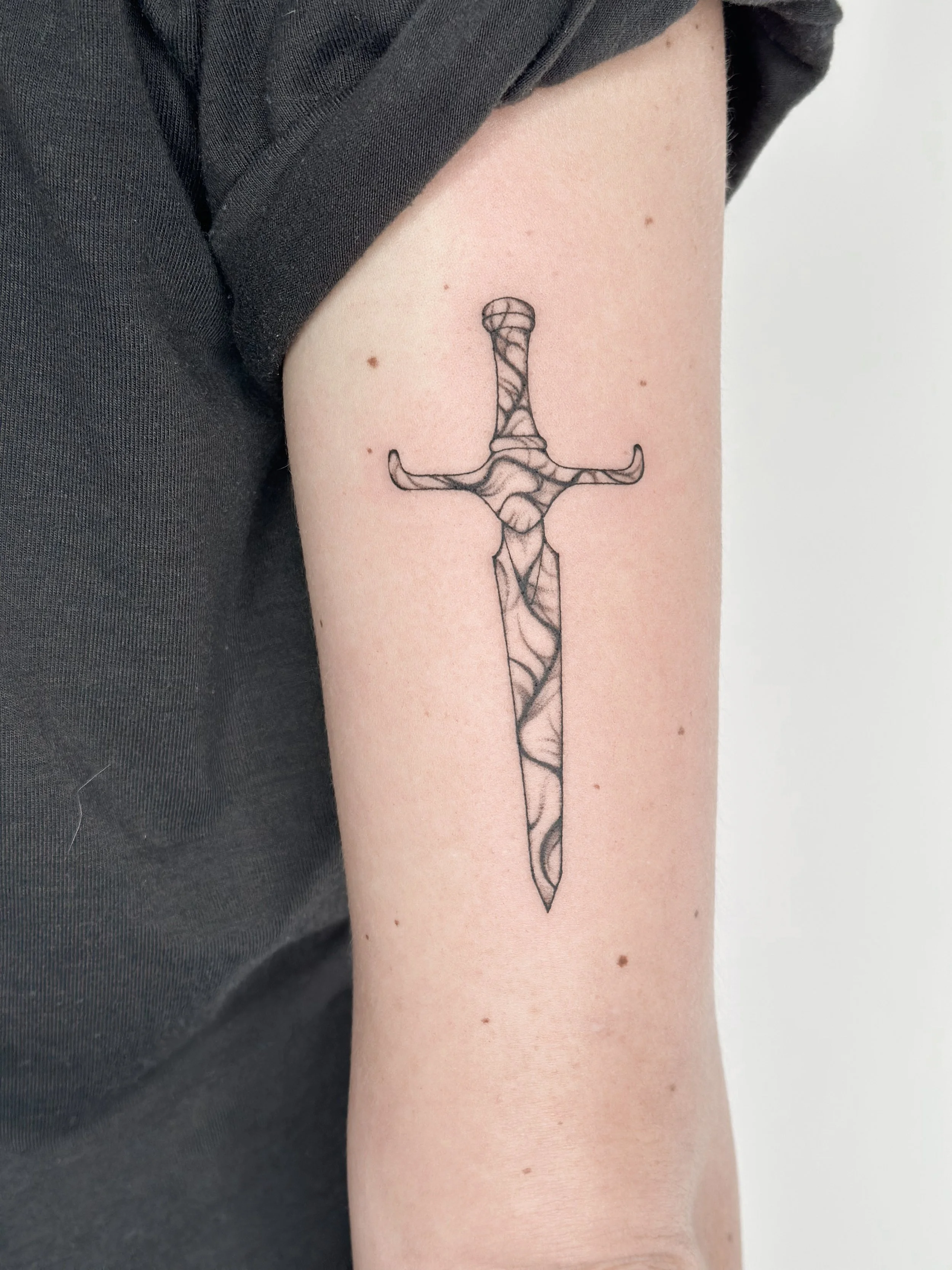 Black outline tattoo of a sword with a wavy, patterned design on a person's upper arm.