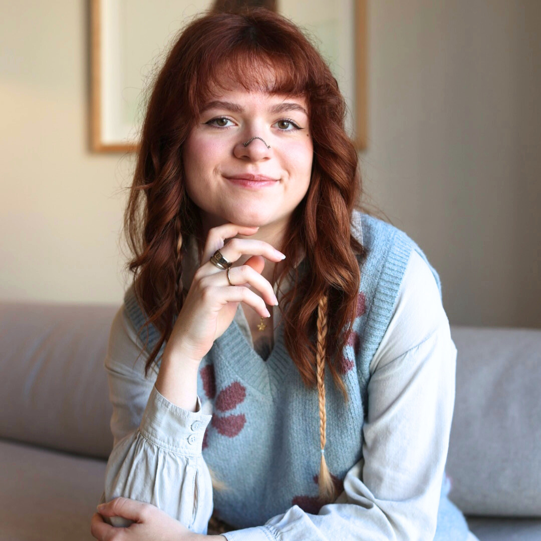 Person with red hair, smiling, wearing a light blue patterned vest over a white blouse, sitting on a couch.