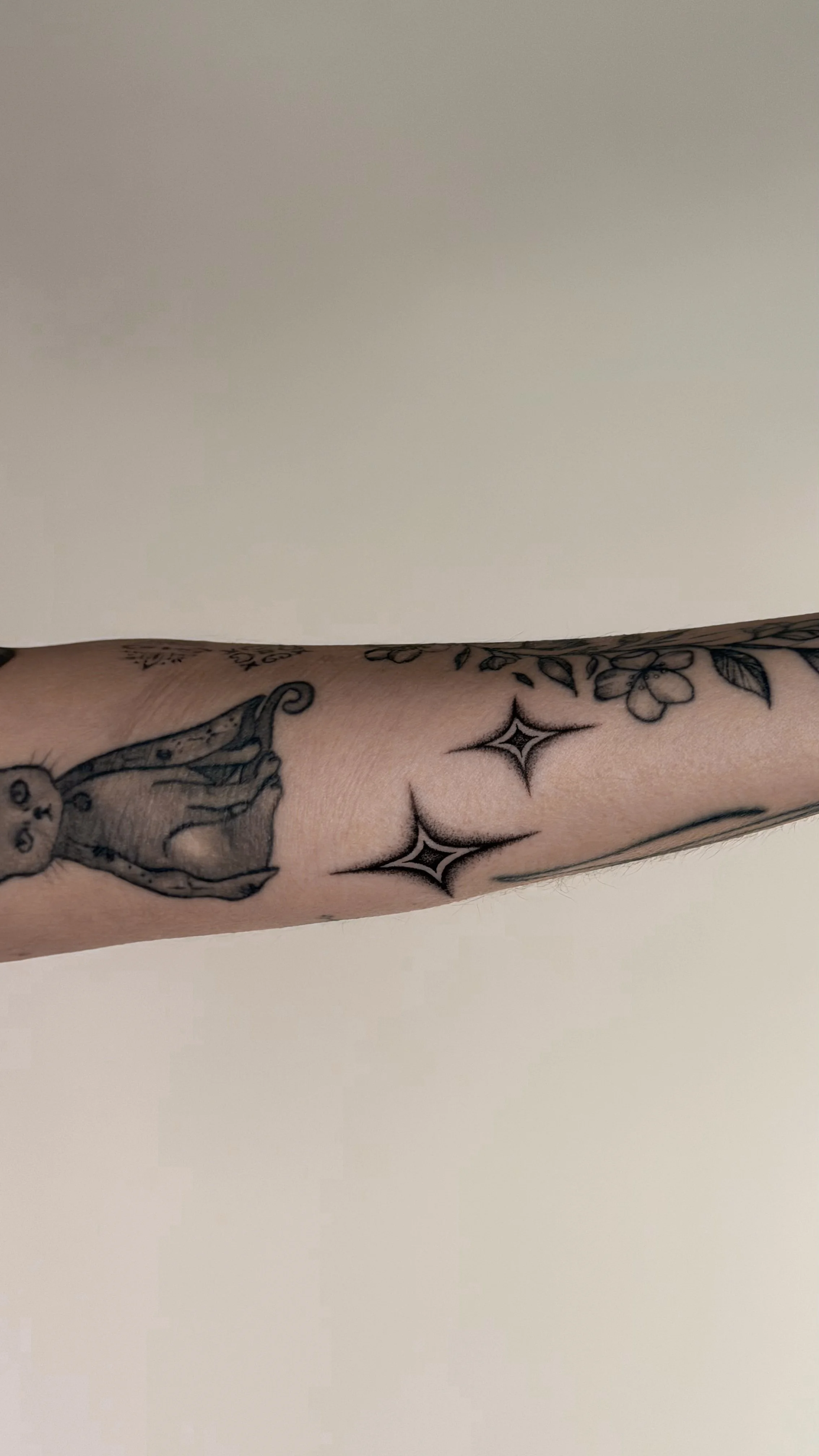 A person's arm with tattoos, including a cartoon cat on the forearm and three star-like designs near the elbow, against a plain neutral background.