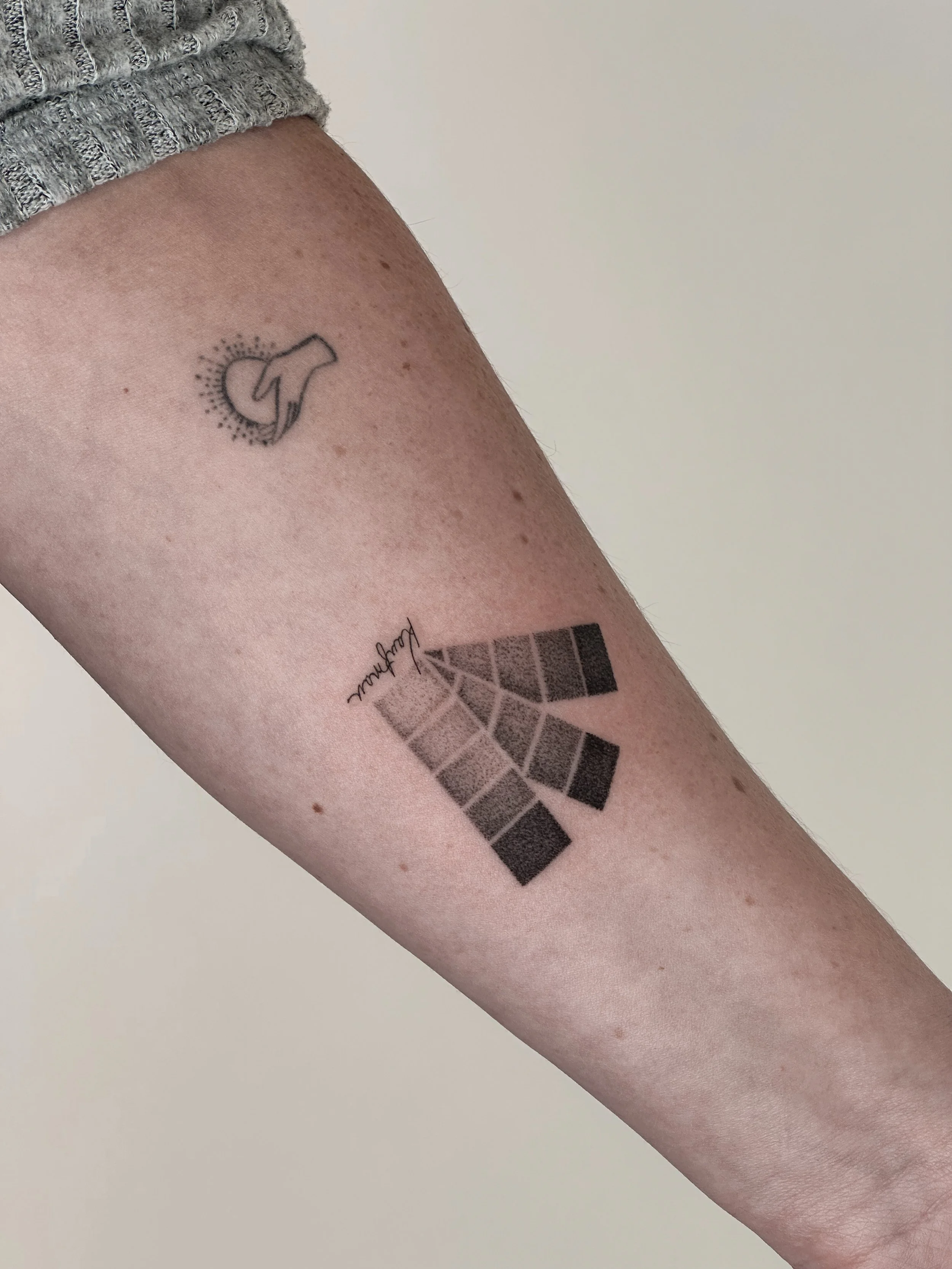 A person's arm with two tattoos: a small line art tattoo of a hand holding a heart surrounded by dots near the shoulder, and a larger geometric and dotwork moth tattoo with the word "growth" written along one wing.