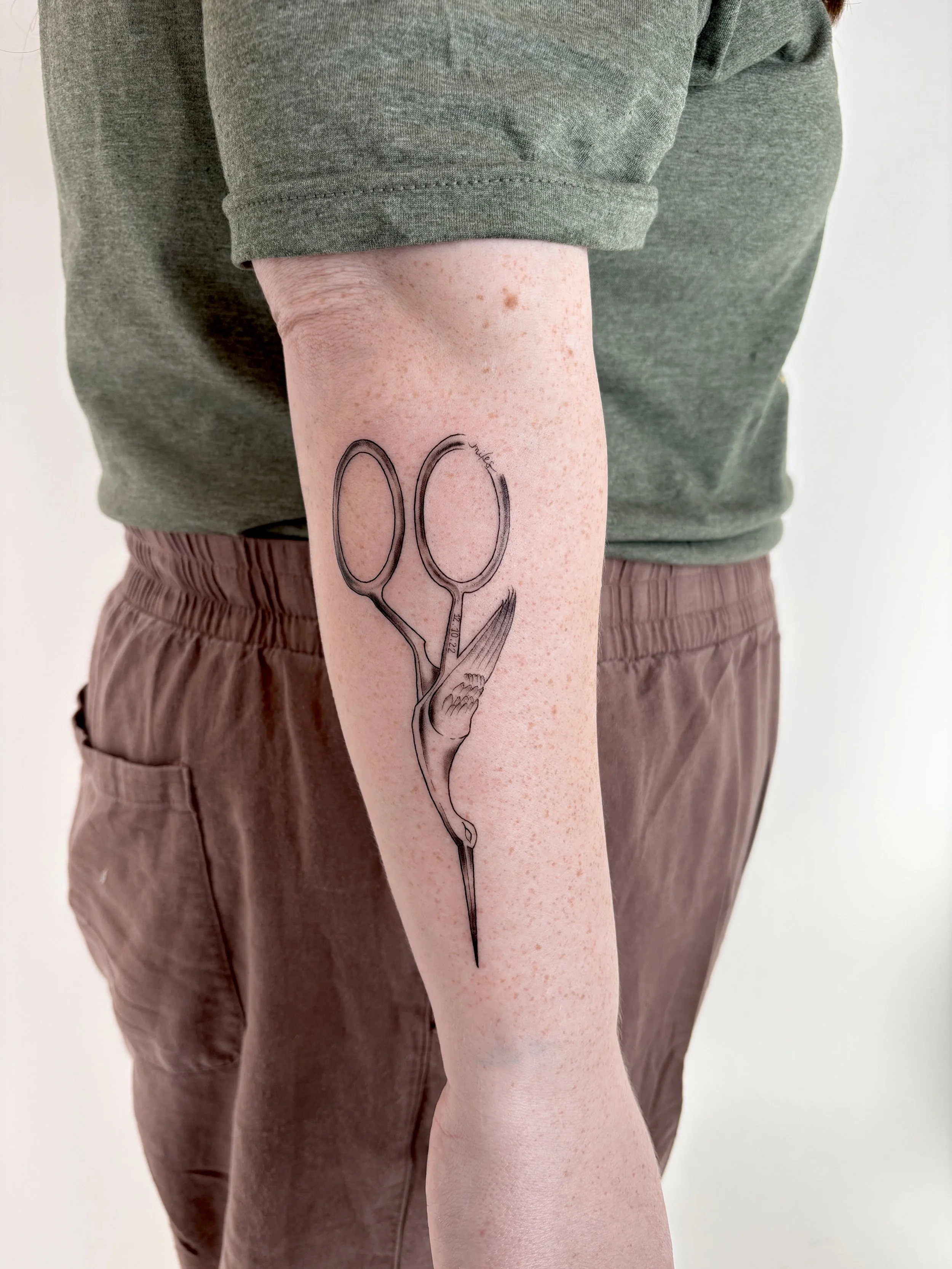Person with a tattoo of stork sewing scissors along their forearm.