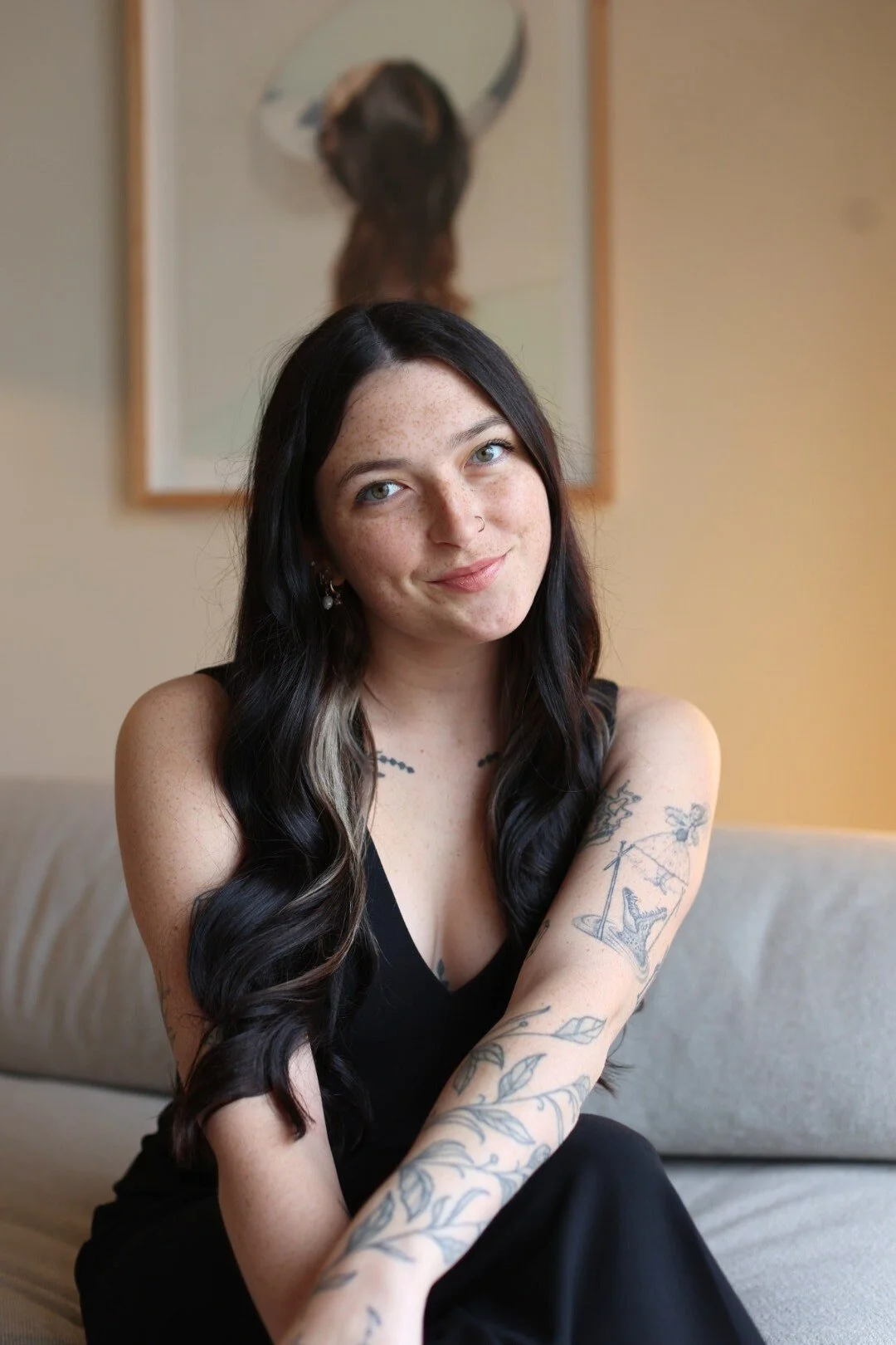 Woman with long dark hair sitting on a couch, wearing a black dress, and showing tattoos on her arms.