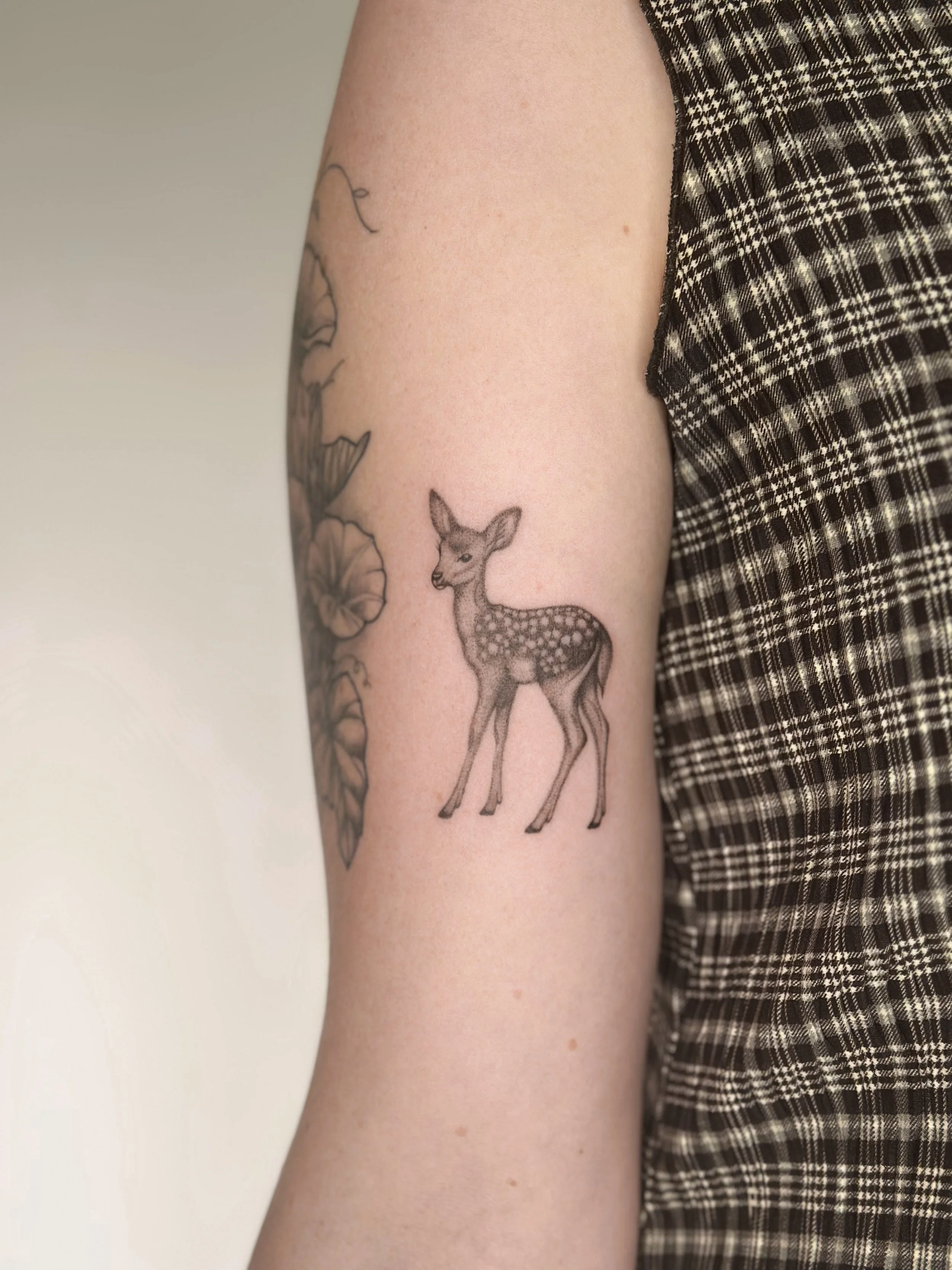 Tattoo of a deer on a person's upper arm, next to a floral design, with the person wearing a plaid sleeveless top.