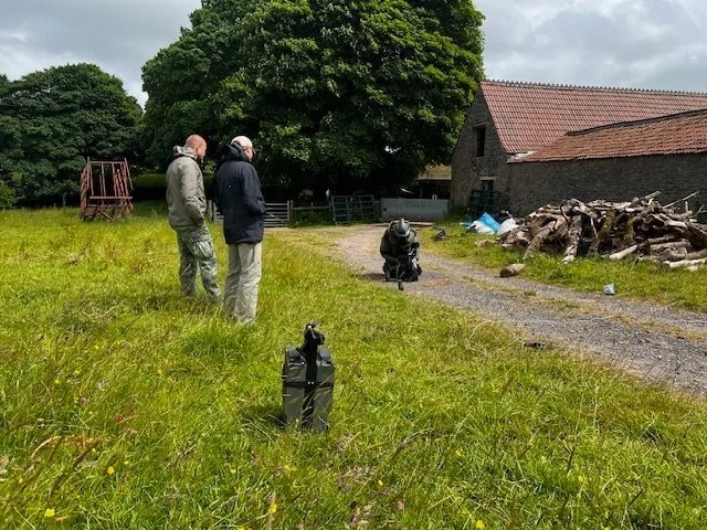 Three men standing outdoors on a grassy field with another person taking a photograph or video. There is a large tree, a building with a red roof, and a pile of wood in the background.