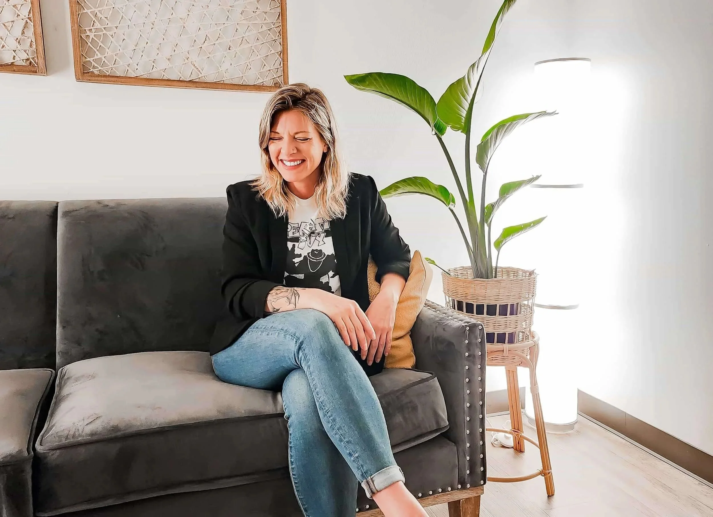 A blonde woman wearing a black jacket and denim pants, sitting on a couch smiling. There is a plant to her left side.