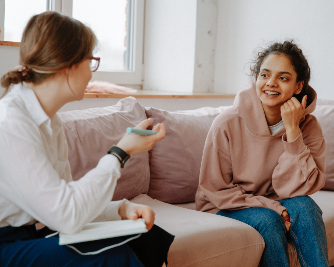 Female therapist sitting on couch performing a therapy session with a female teen client wearing a pink sweater.