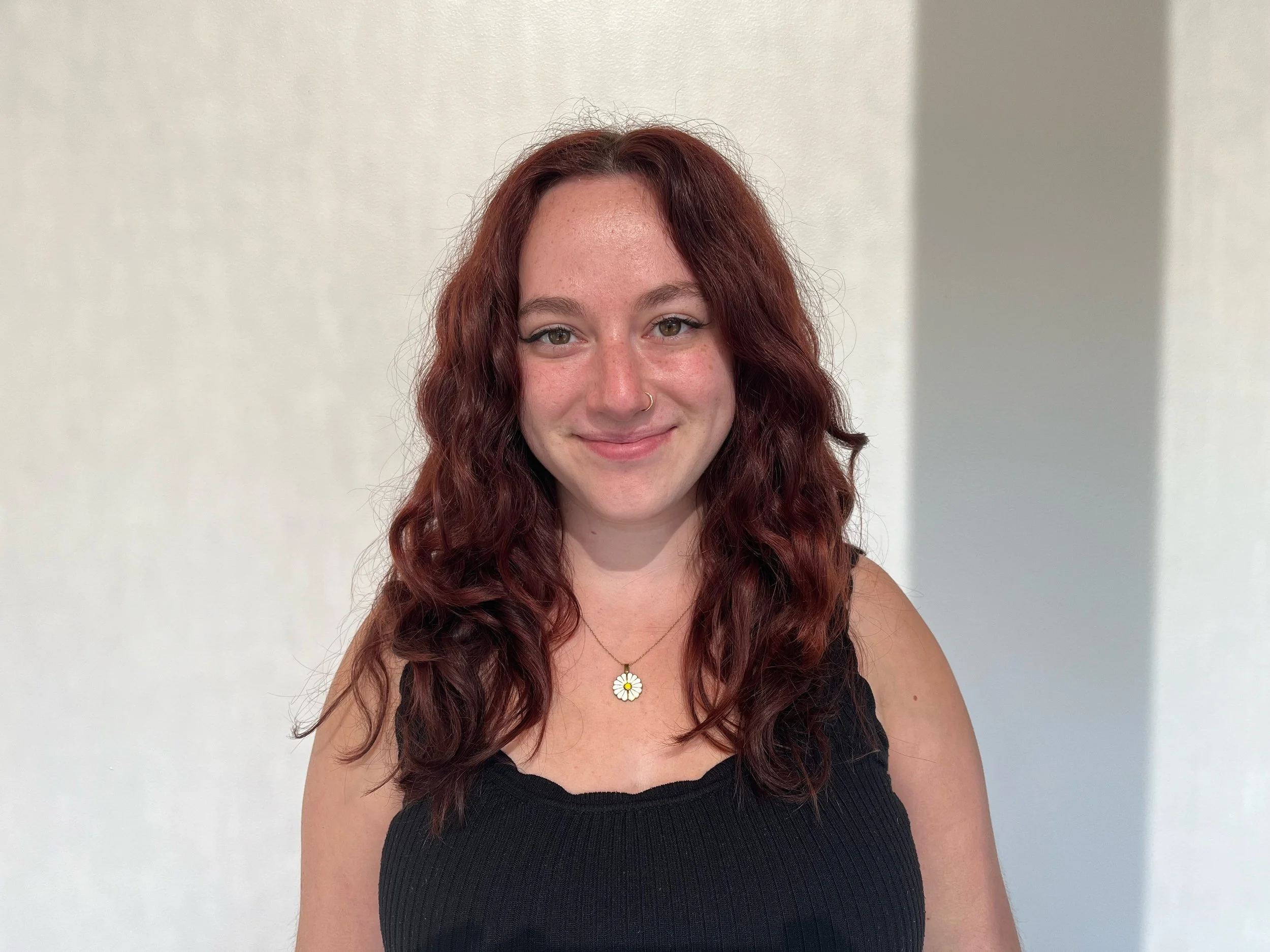 A young woman with shoulder-length wavy red hair, wearing a black sleeveless top and a daisy-shaped necklace, smiling in front of a plain off-white wall.