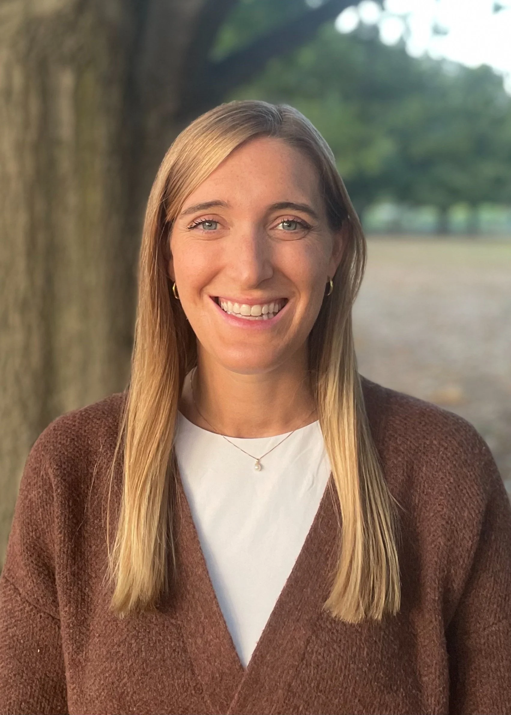 Woman smiling outdoors with long blonde hair, wearing a brown cardigan and white top, standing in front of tree and blurred background.