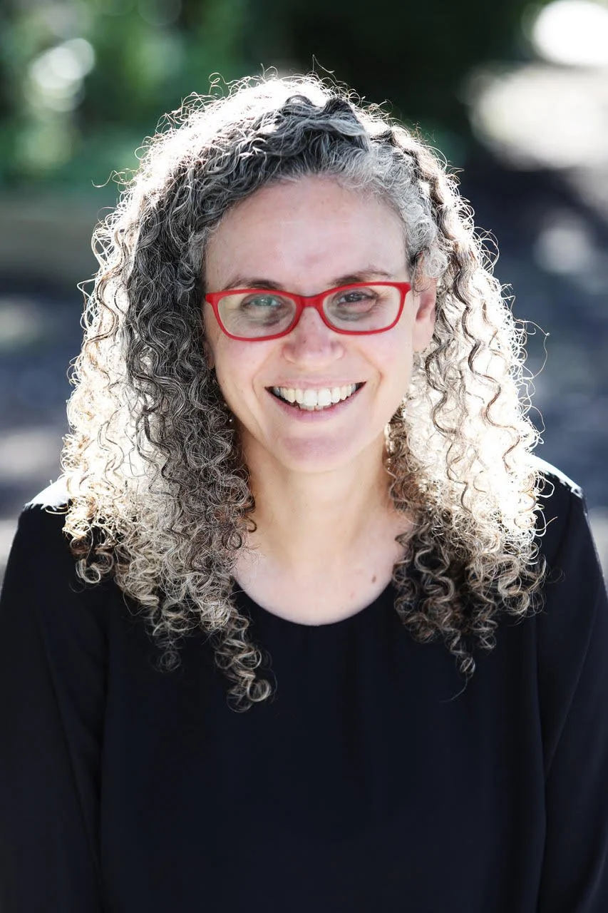 Person with curly gray hair and red glasses smiling outdoors