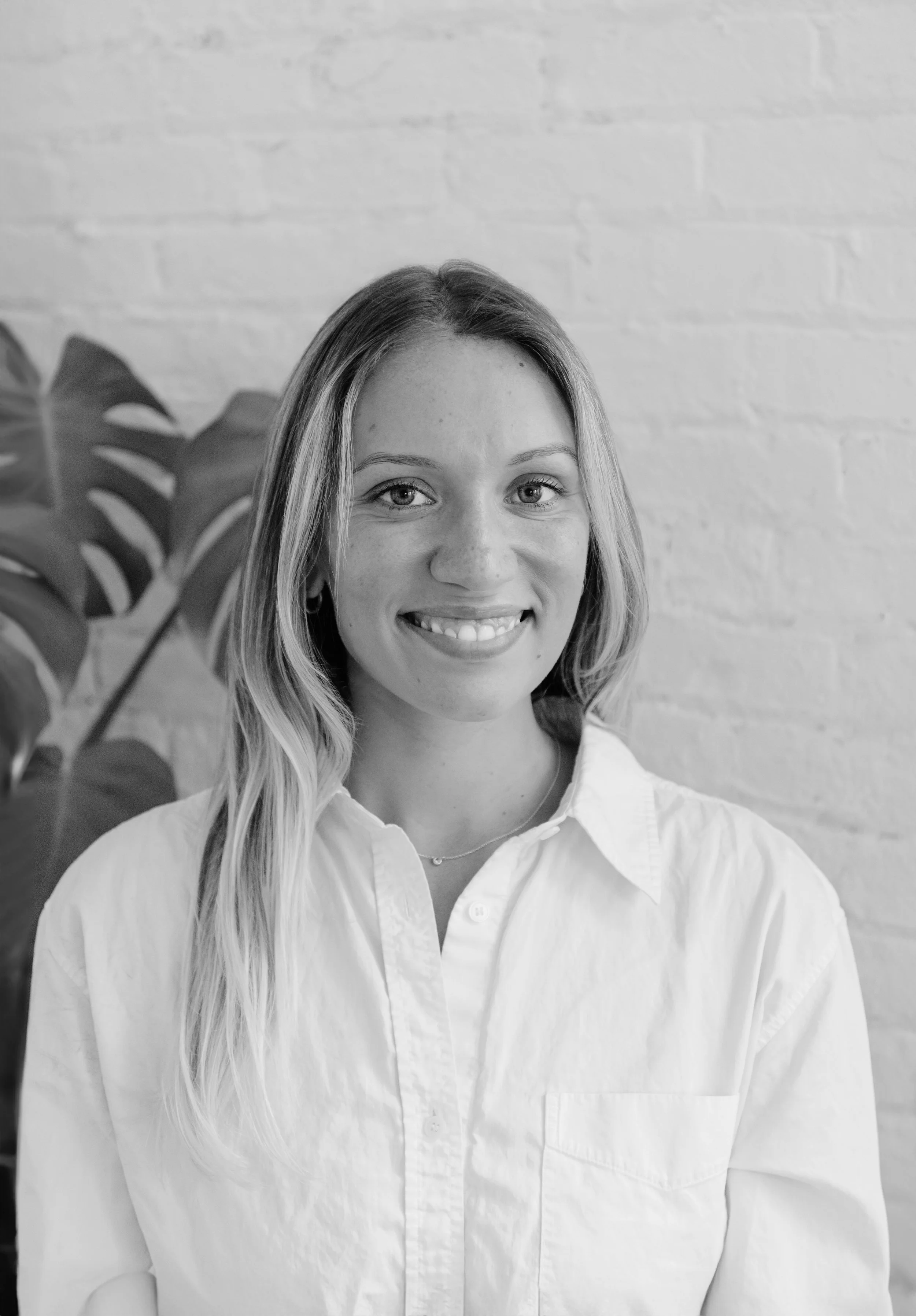 A young woman with long, wavy hair smiling at the camera, wearing a white button-up shirt, with a plant and a brick wall in the background.