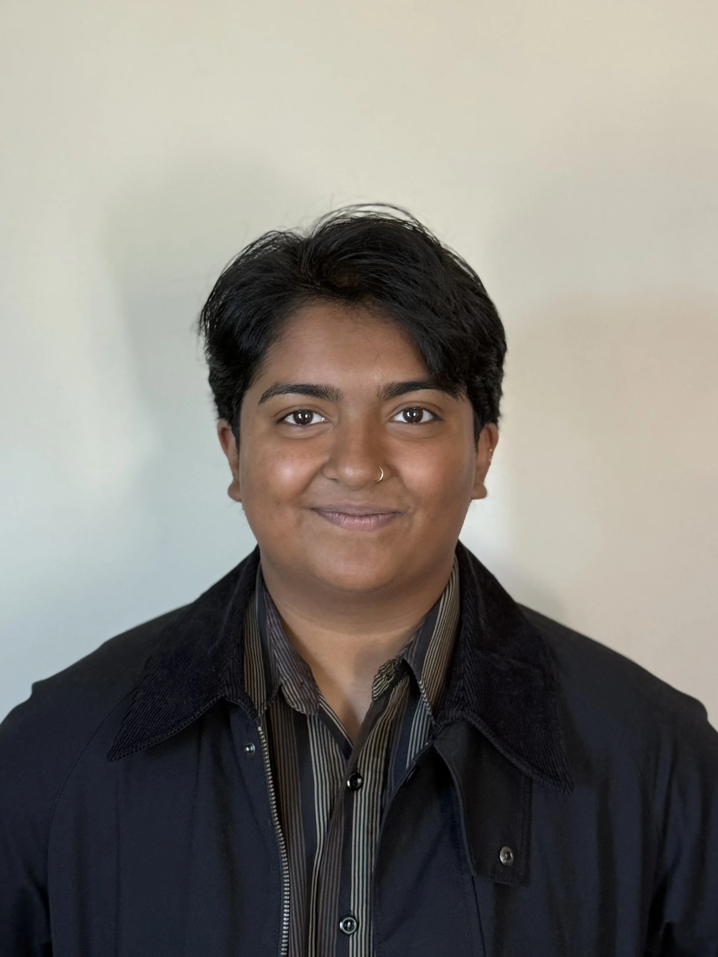 Headshot of a person with short dark hair, wearing a black jacket over a striped shirt, smiling slightly, in front of a plain light-colored background.