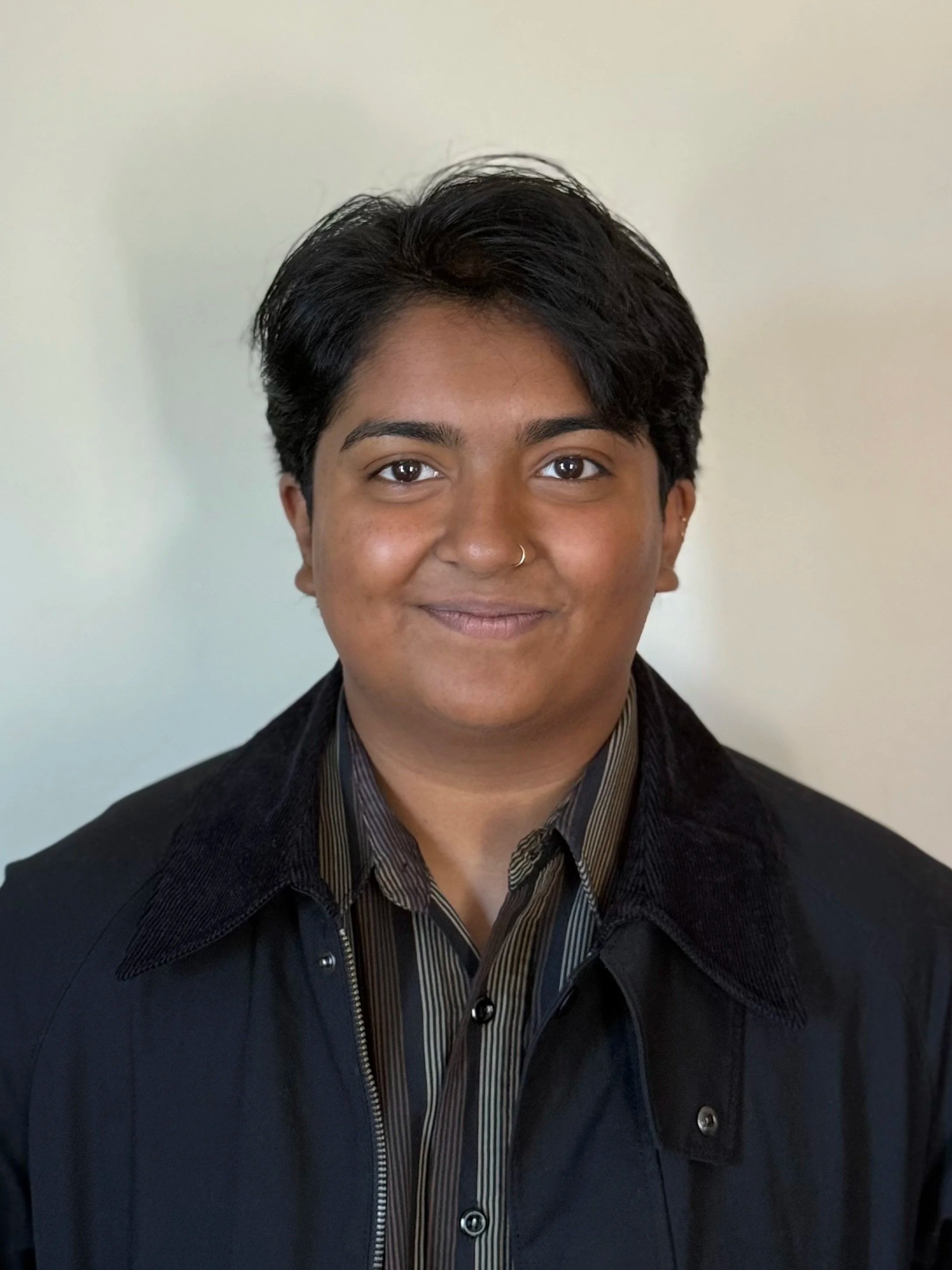 Portrait of a person with short dark hair, wearing a black jacket over a striped shirt, against a plain, light-colored background.