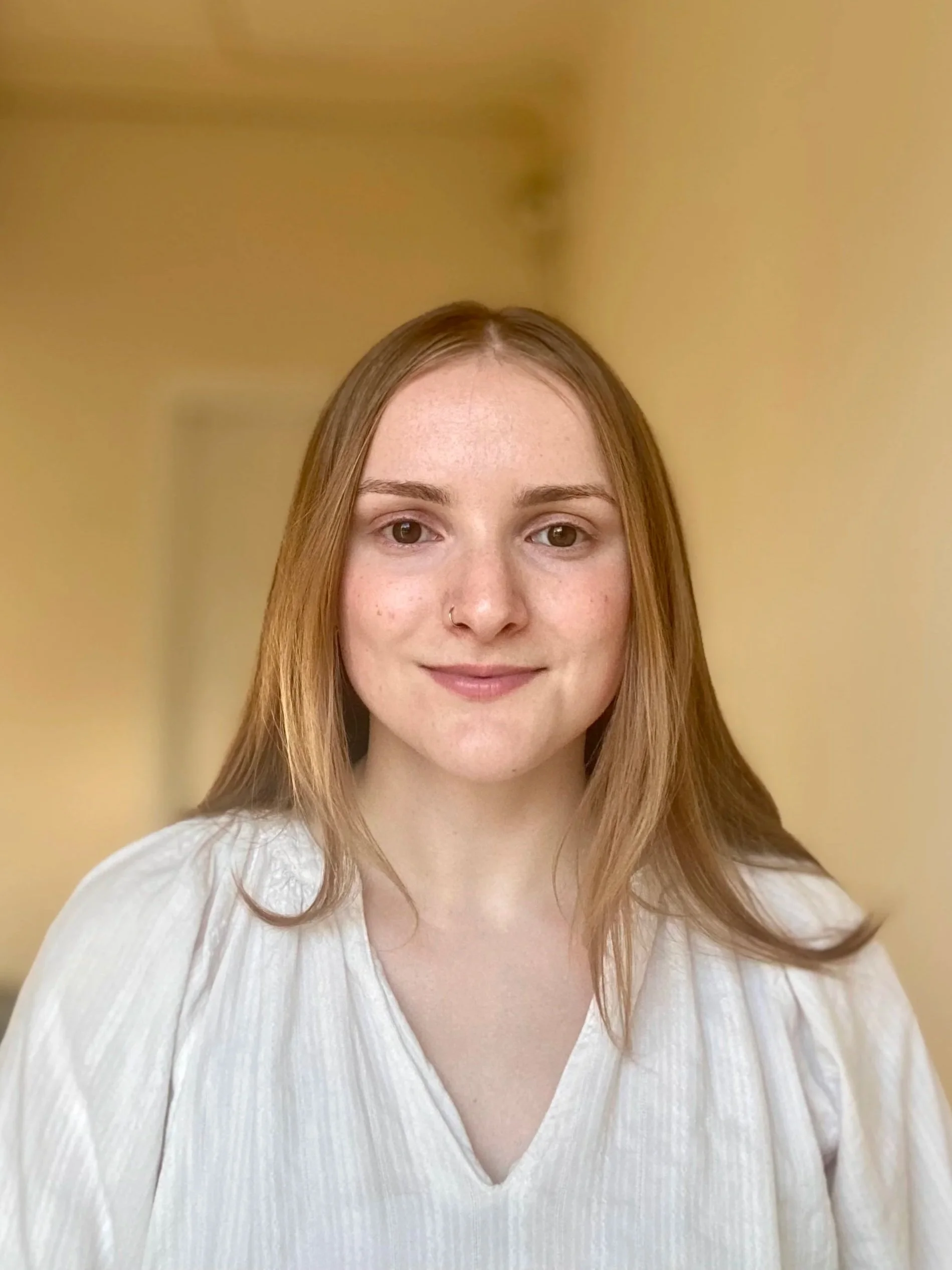 A young woman with red hair and a nose piercing smiling in a white top, standing indoors with a yellow wall background.