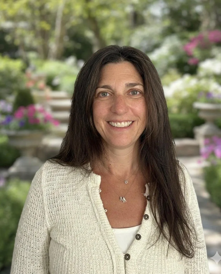 A woman with long dark hair smiling, wearing a white cardigan and jewelry, standing outdoors with blurred background of greenery and colorful flowers.