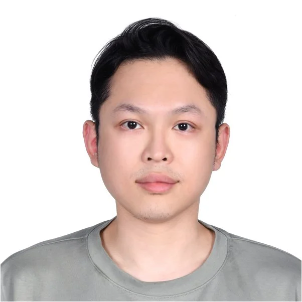 A portrait of a young Asian man with black hair, wearing a gray shirt, looking directly at the camera against a white background.