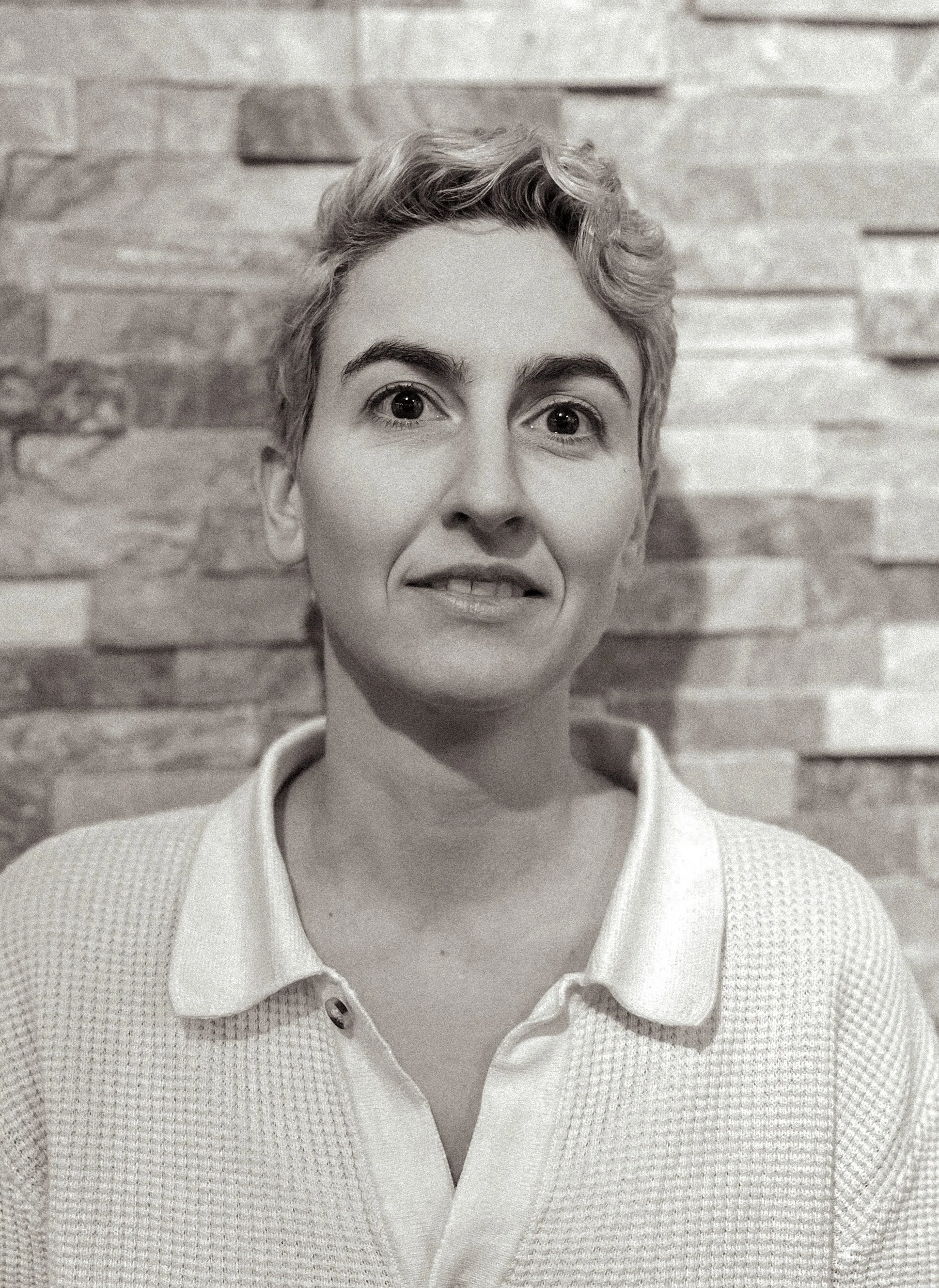 A woman with short, curly hair and a collared shirt, standing in front of a textured brick wall.