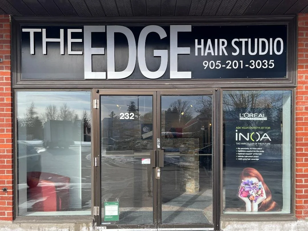 Exterior front view of 'The Edge Hair Studio' with glass doors and windows, showing the street and trees reflected in the glass, and a large sign with the salon name, contact number, and promotional poster about hair coloring services.