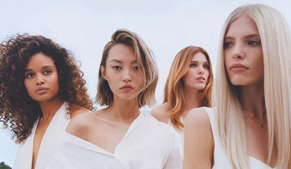 Four women standing outdoors with serious expressions, wearing white tops against a cloudy sky background.