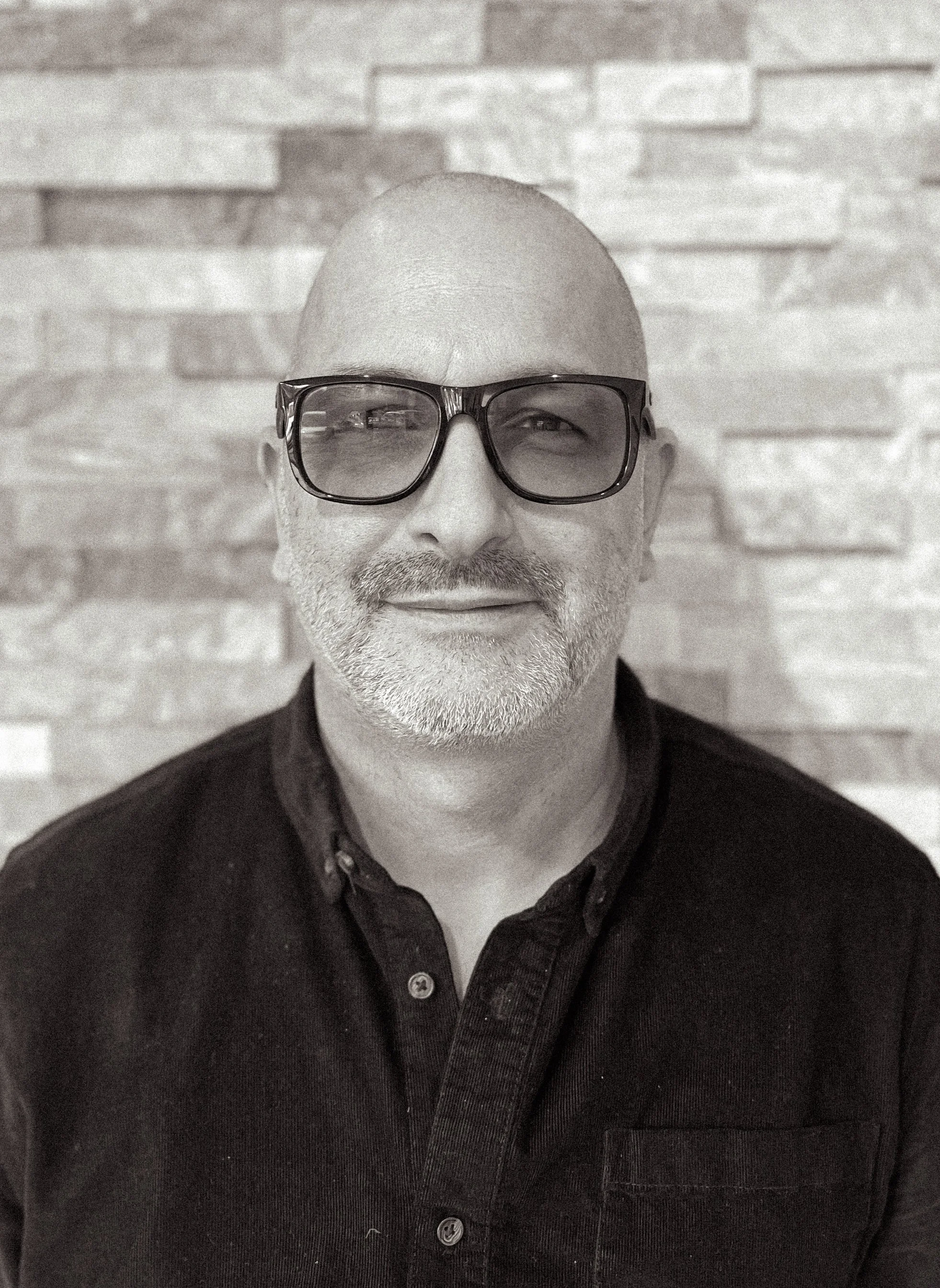 A middle-aged man with a bald head and gray beard, wearing large dark glasses and a dark button-up shirt, smiling in front of a textured brick wall.