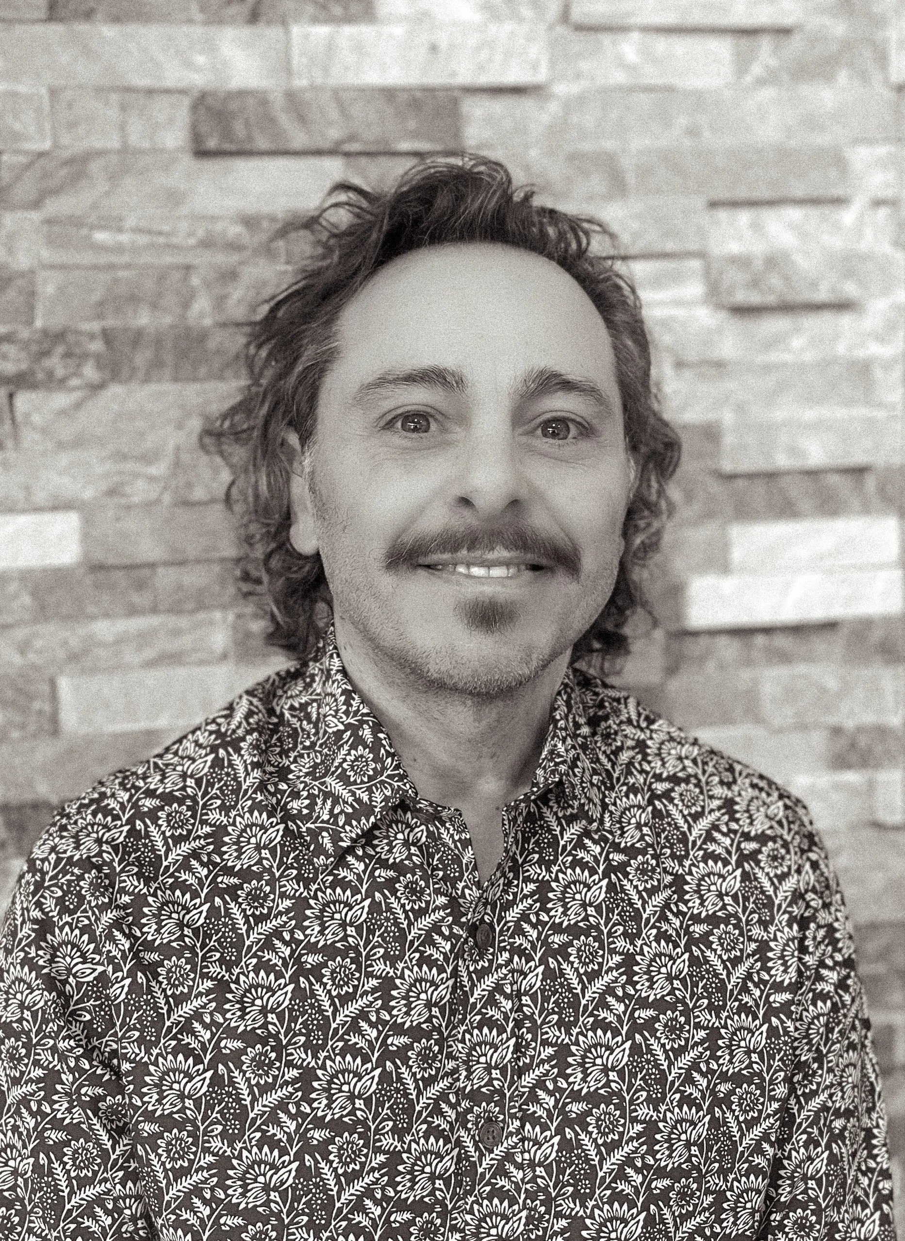 Black and white portrait of a man with curly hair, a mustache, and goatee, wearing a floral patterned shirt, smiling, with a brick wall background.