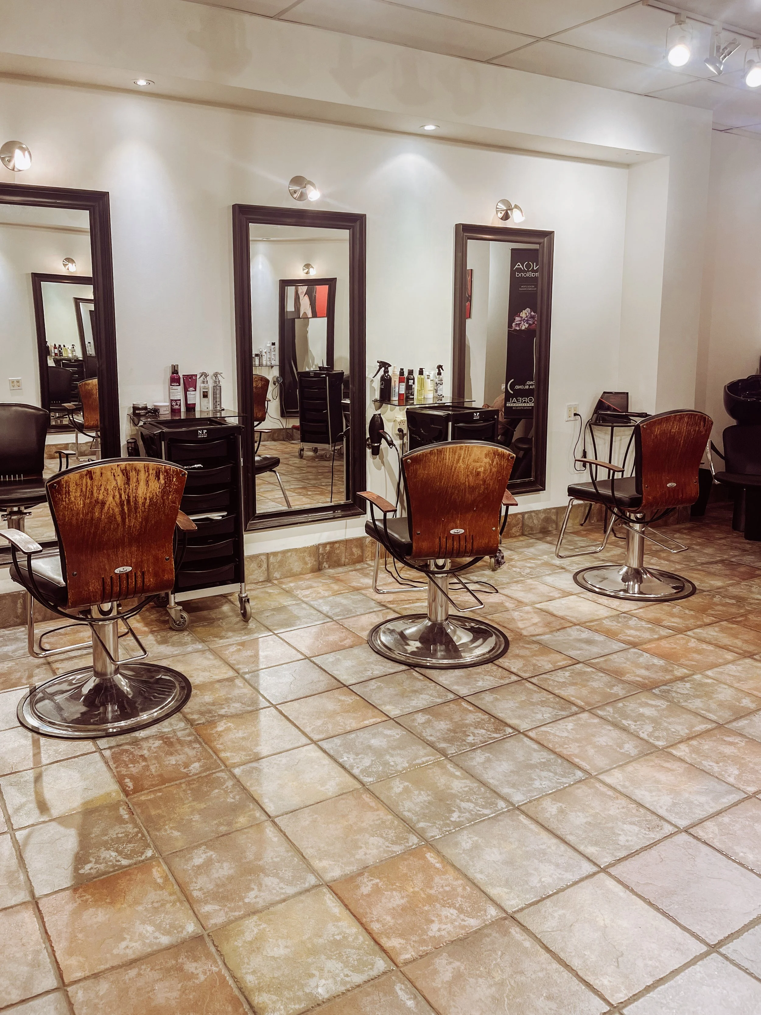 Inside a hair salon with three styling stations, each featuring a mirror, chair, and on-wheeled trolleys with hair products and tools. The salon has tiled flooring and ceiling lights.