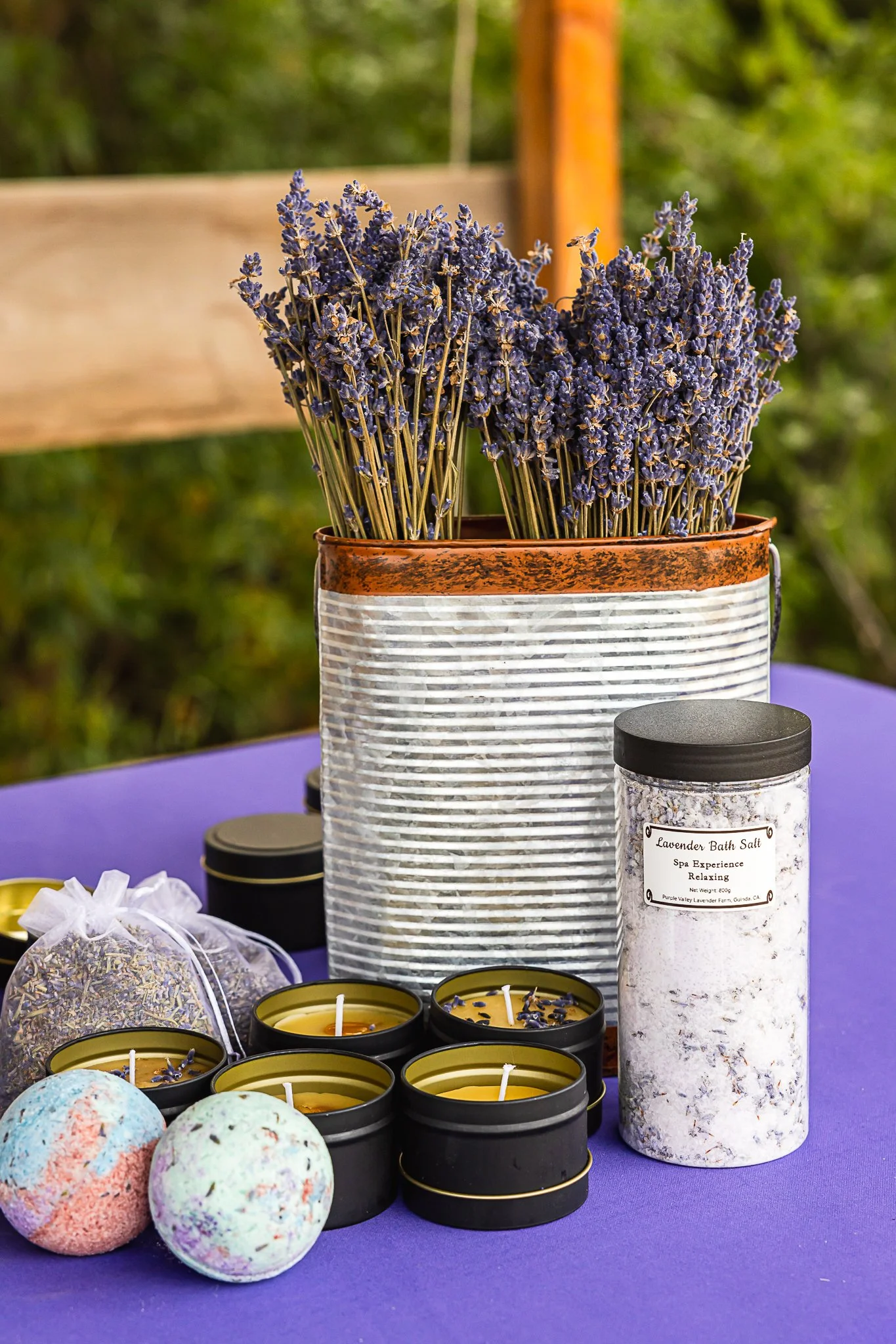 Dried lavender stems in a metal container surrounded by small candles, bath bombs, and a jar labeled 'Lavender Bath Salt' on a purple table.