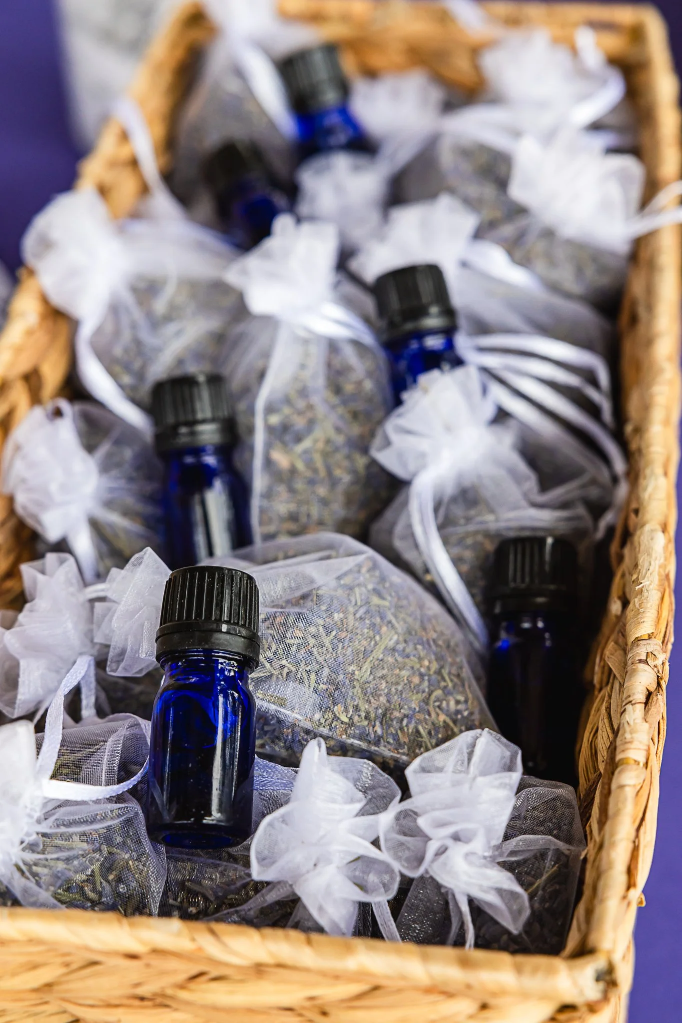 A wicker basket containing small blue glass bottles with black caps surrounded by white organza bags filled with dried lavender.