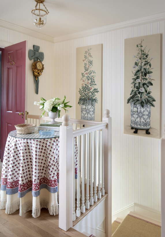 Interior of a cozy room corner with cream striped wall panels, a pink door, and vintage decor. A round table with a red, white, and blue patterned tablecloth holds a flower arrangement and some books. Two botanical art prints hang on the wall, and a 