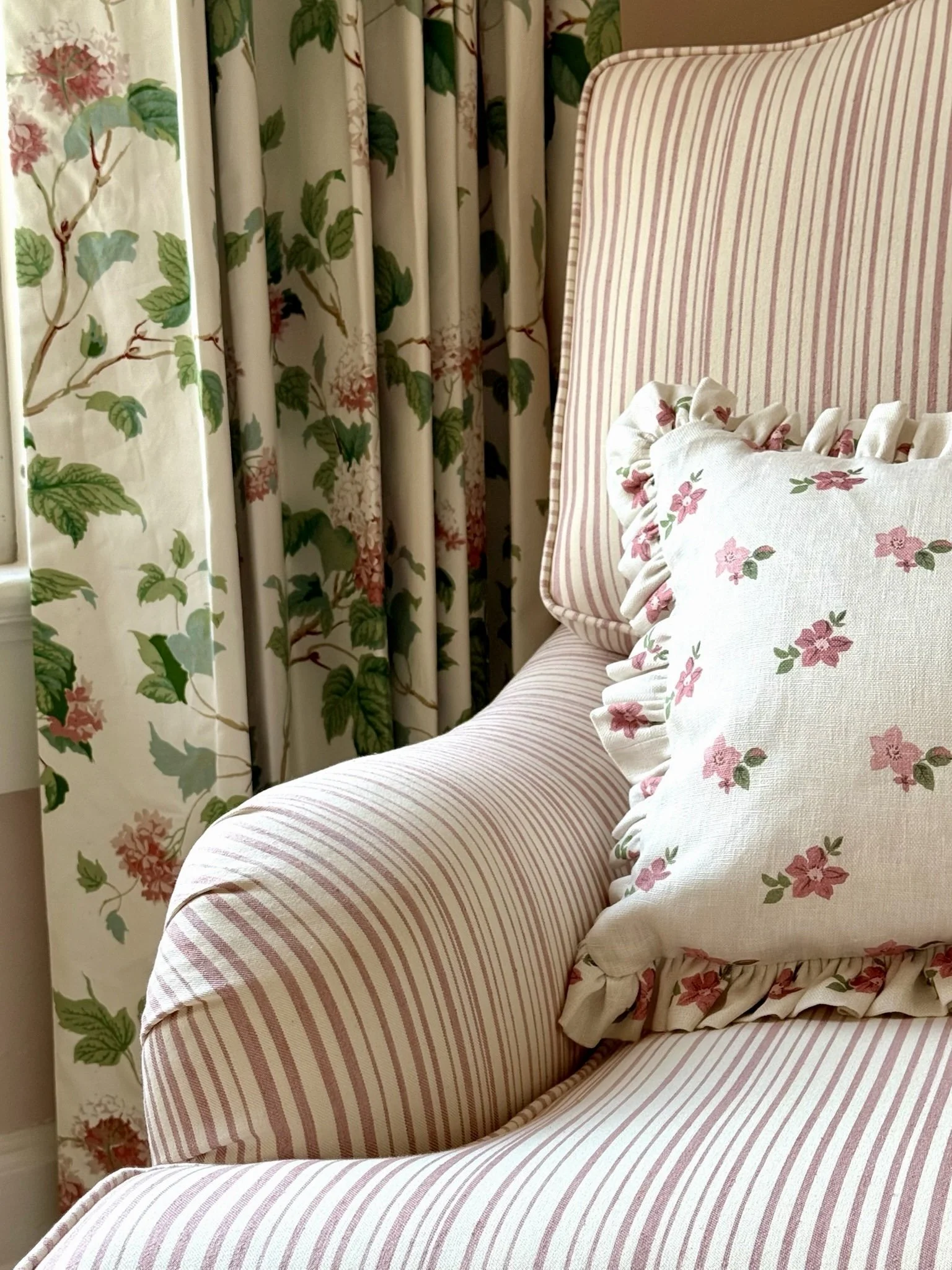 Close-up of a pink and cream striped armchair with ruffled floral pillow, with floral patterned curtains in the background.
