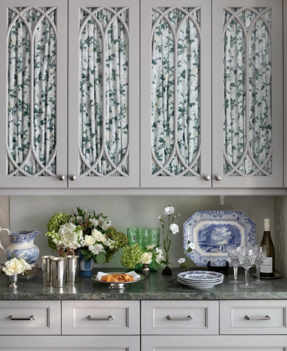 A kitchen countertop decorated with flowers, dishes, and glassware, with cabinets featuring patterned curtains.