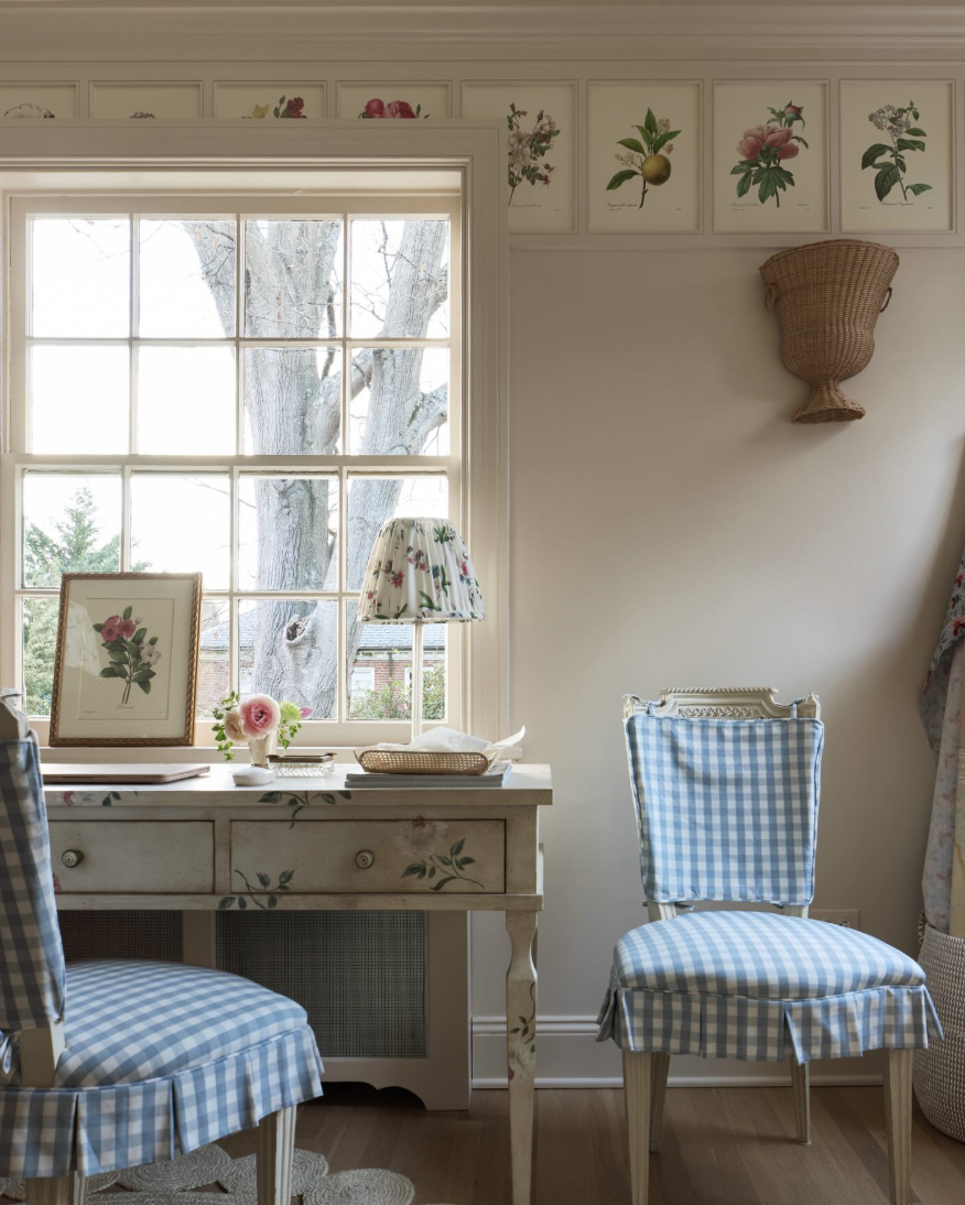 A vintage-style room with a window showing tree branches outside. On the wall, there are framed botanical prints and a wicker wall hanging. A distressed white desk with floral designs holds a framed flower print, a small vase with a pink flower, and 