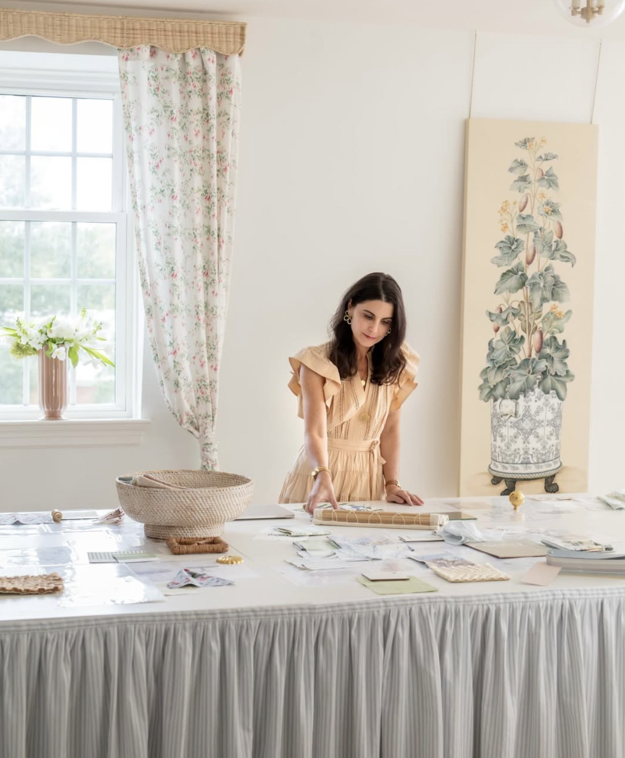 Table covered with fabric swatches, papers, and design materials, in a bright room with white walls, floral curtains, a window with greenery outside, and tall artwork designed by Richmond, VA interior designer Jenny Holladay Design.