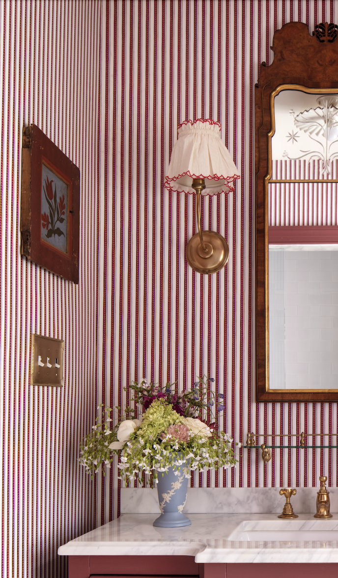 Close-up of a vintage-style bathroom wall with red and white striped wallpaper, a framed floral artwork, a wall-mounted brass lamp with a fabric shade, a mirror with an ornate wooden frame, a blue vase with a floral arrangement on a marble countertop