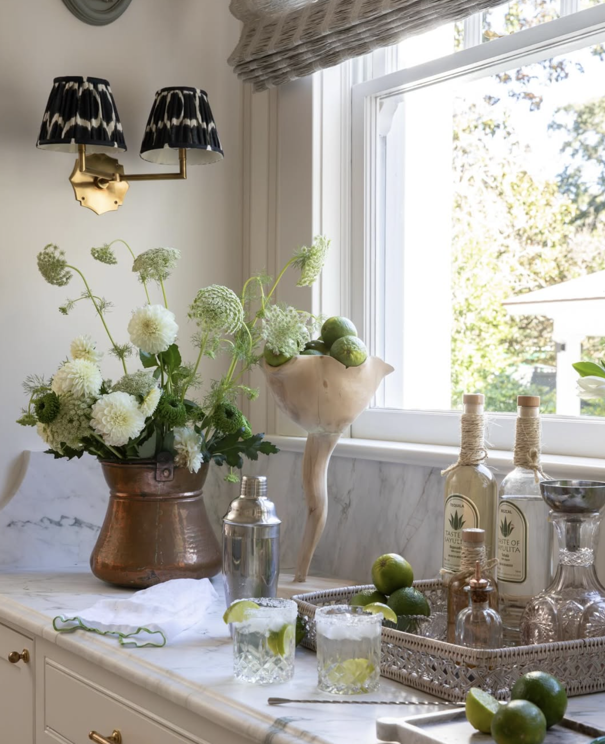 A kitchen counter with a flower arrangement, a bowl with limes, bottles with twine, a metallic shaker, glasses with lime slices, and a tray with limes, near a window with a view of trees.