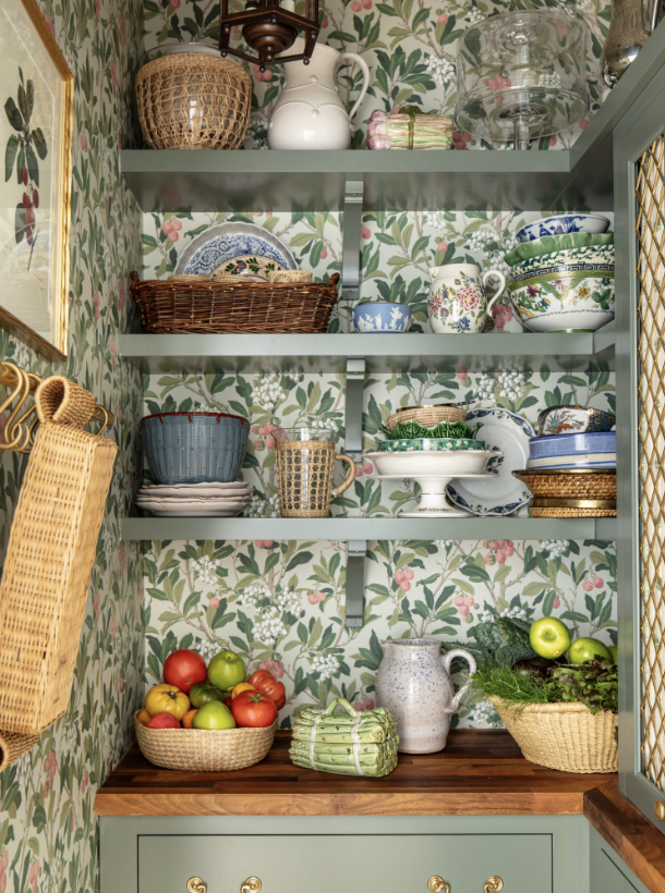 Kitchen hutch with green floral wallpaper, displaying various ceramic dishes, bowls, and pitchers, with baskets of apples and leafy greens on a wooden countertop created by Jenny Holladay Design in Richmond, VA.