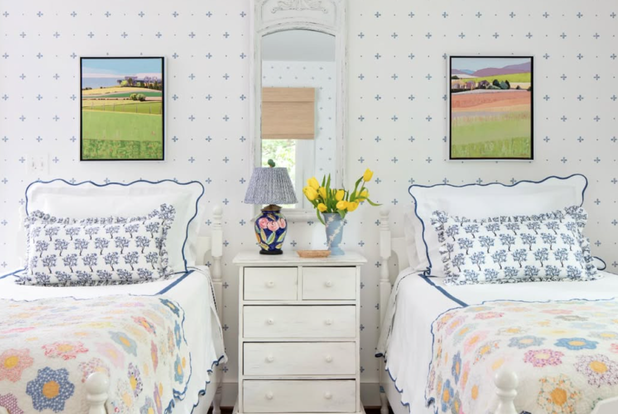 A cozy bedroom with two white twin beds with blue trim, floral pillows, and pastel quilts. A white nightstand with a colorful vase of yellow tulips, a decorative lamp, and a small tray is between the beds. Above the beds are two landscape paintings. 