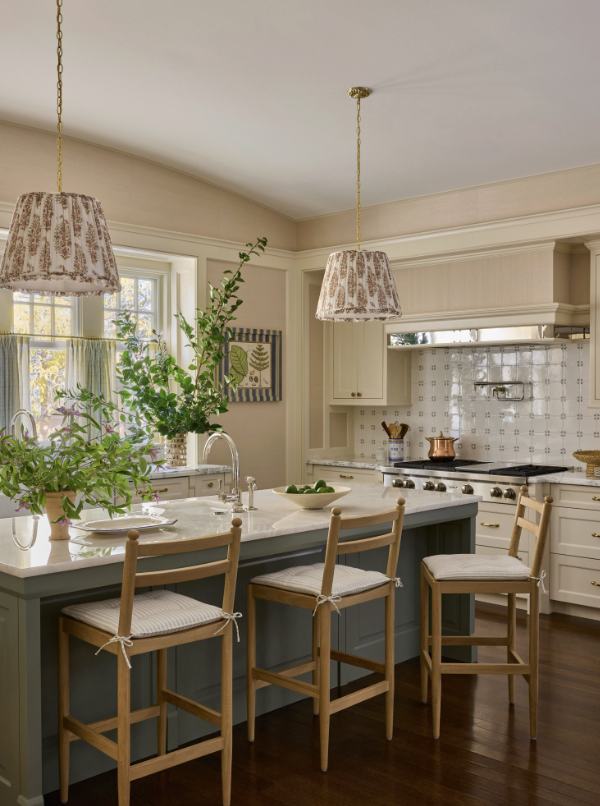Bright kitchen with white cabinetry, a marble island with three wooden chairs, pendant lights, and a window with sunlight, greenery, and artwork created by Jenny Holladay Design in Richmond, vA 