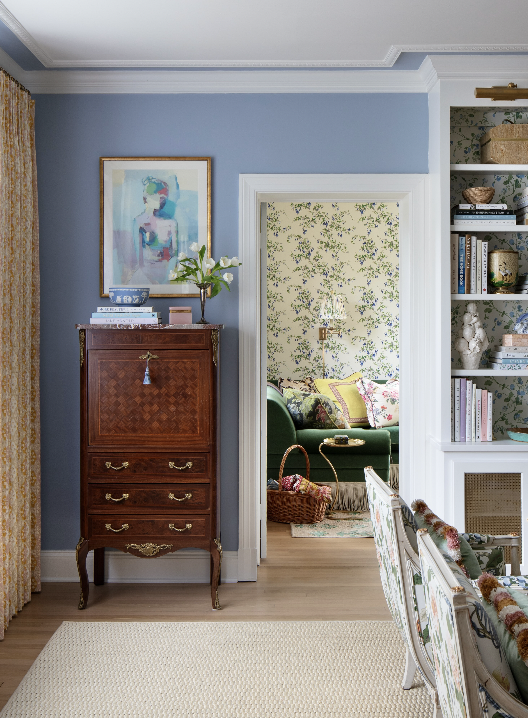 Living room with blue painted walls, a wooden dresser with a vase of flowers and books, and a doorway leading to a room with floral wallpaper and a green sofa.