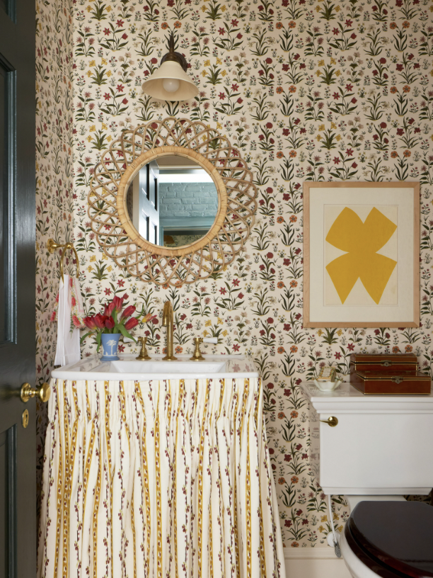 A vintage bathroom with floral wallpaper, a round wicker mirror, a white sink with a gold faucet and a skirt made of fabric with yellow and red patterns, a white cabinet with decorative boxes created by Jenny Holladay Design in Richmond, VA.