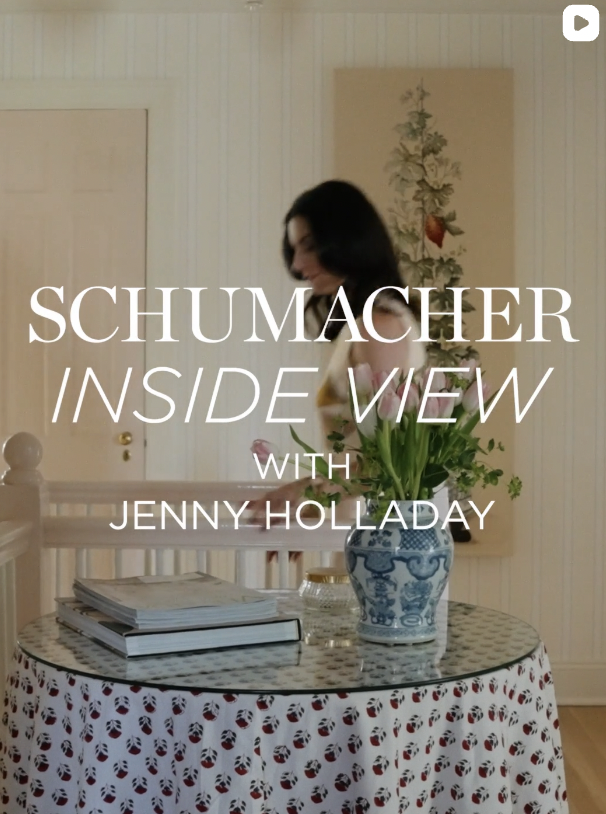 A woman with black hair standing behind a round table with a patterned tablecloth, a blue and white vase with green plants, a stack of magazines, and a small dish. The background features wood-paneled walls and a framed floral artwork.