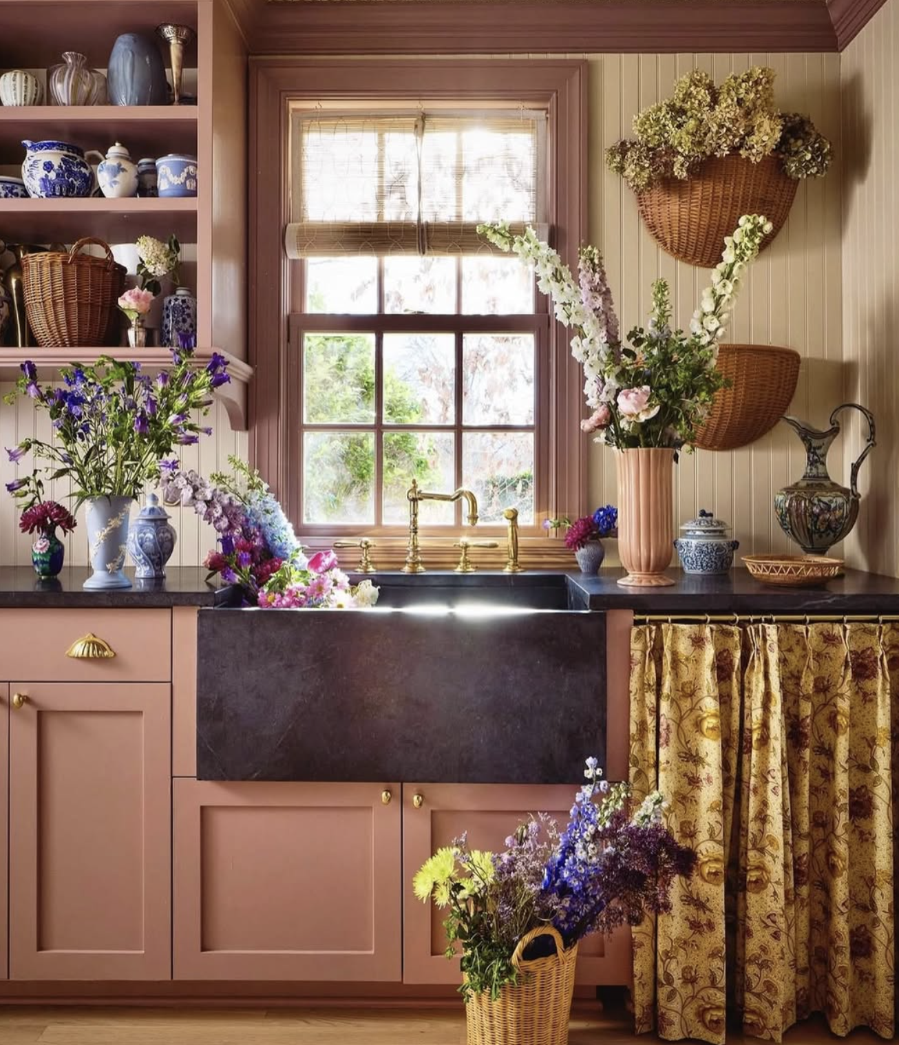 A cozy kitchen scene with pink cabinets, black countertop, and open shelves filled with decorative jars and vases. There are multiple flower arrangements in vases and baskets, a window with sunlight streaming in, and patterned curtains.