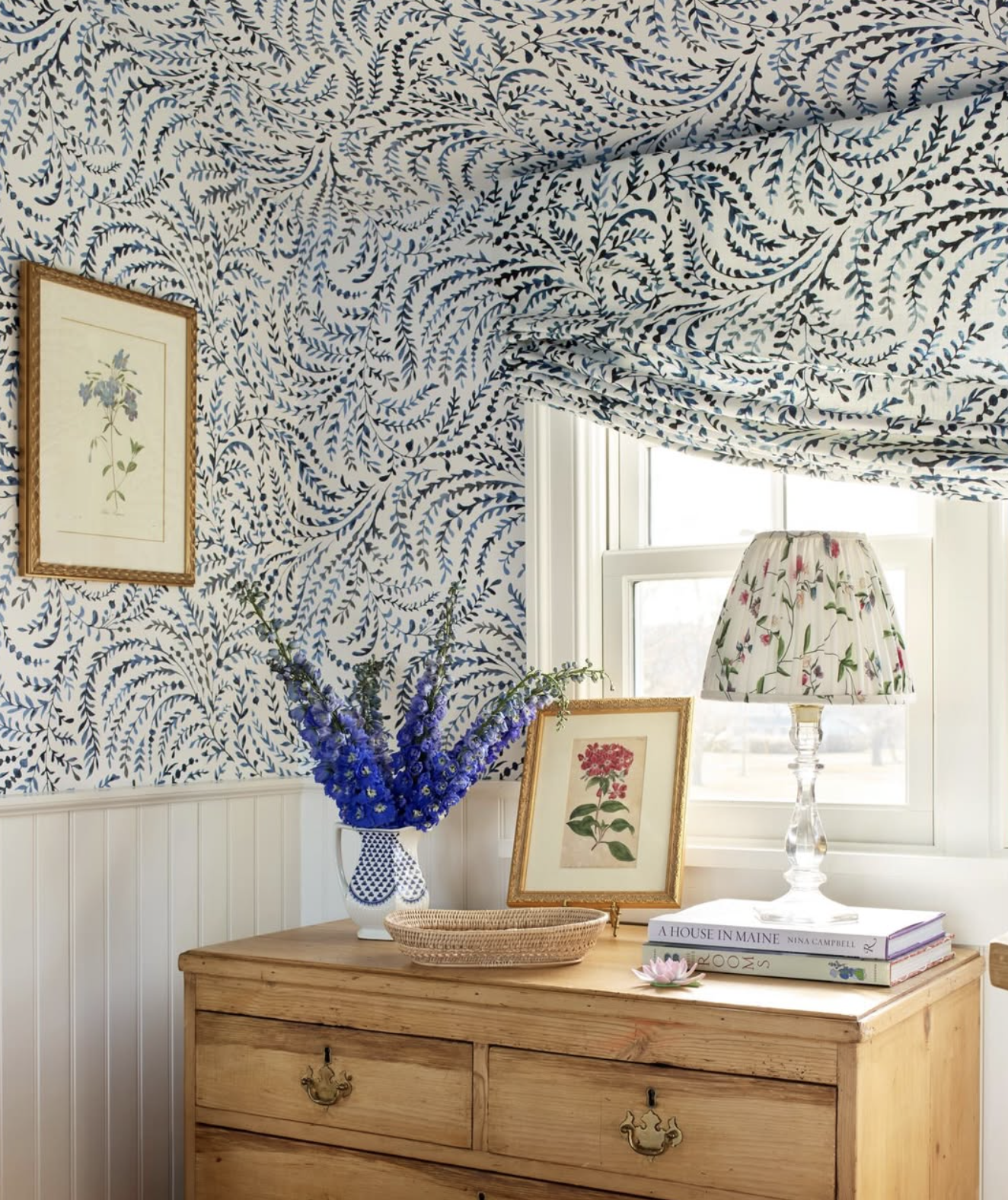 A wooden dresser with a framed floral print, a vase with purple flowers, a framed floral picture, and a floral lamp shade, with patterned wallpaper and a window in the background designed by Richmond, VA interior designer Jenny Holladay Design.
