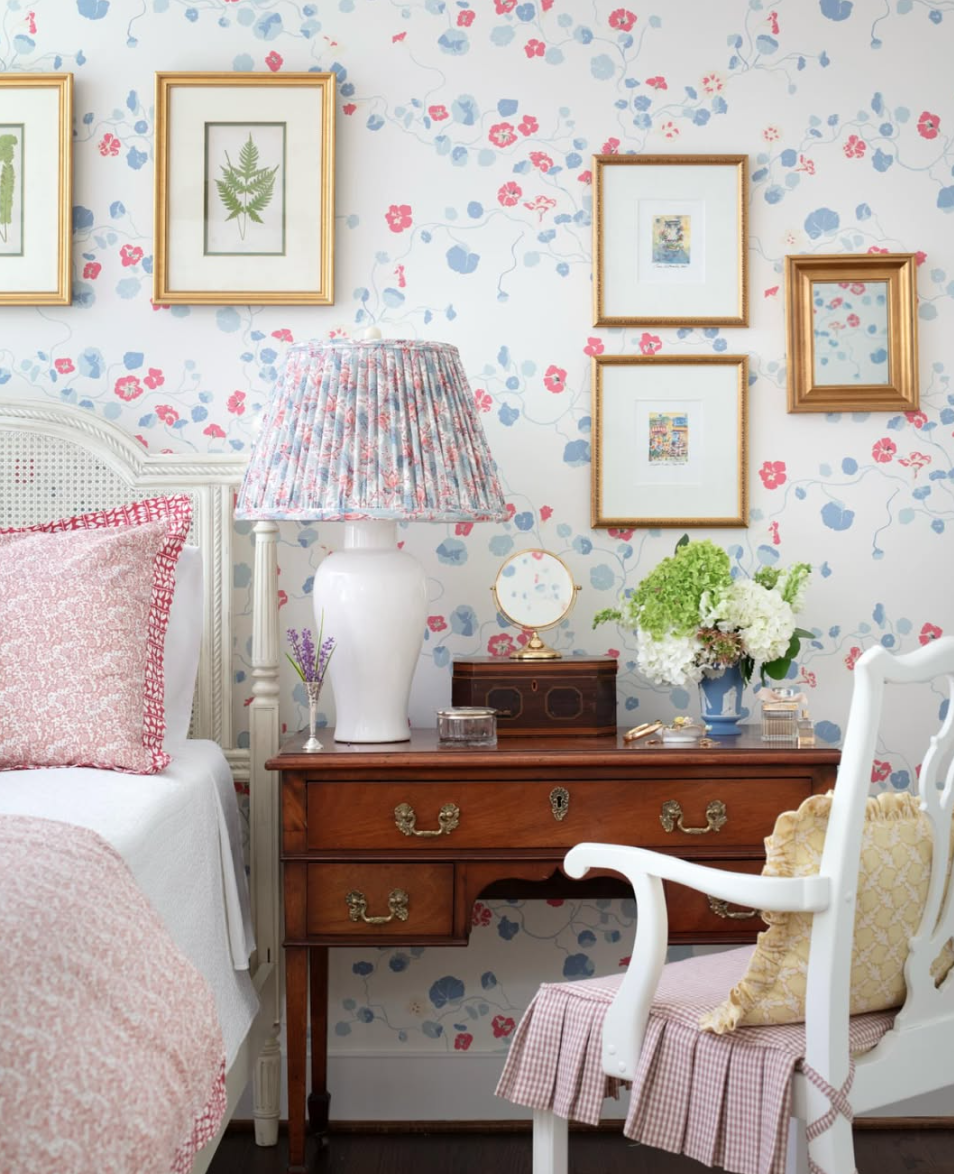 A bedroom corner with a vintage wooden dresser, floral wallpaper, and framed botanical art. The dresser holds a large white ceramic lamp with a floral shade, a small mirror, bedding, and a bouquet of white and green flowers. A white armchair with pat