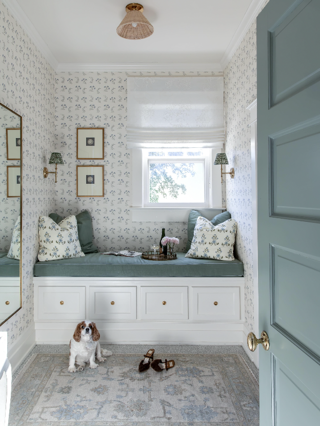 Cozy window seat nook with floral wallpaper, decorative pillows, a tray with flowers, and a small dog sitting on an area rug, seen through an open teal door.