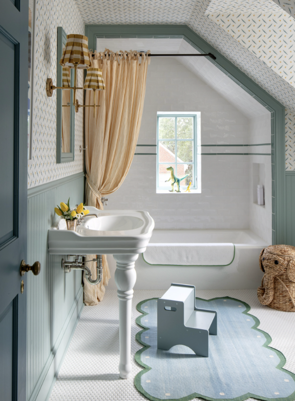 A cozy bathroom with a vintage-style white pedestal sink, a window with a small dinosaur toy on the sill, and a bathtub with a blue and green scalloped-shaped rug in front created by Jenny Holladay Design in Richmond, VA.