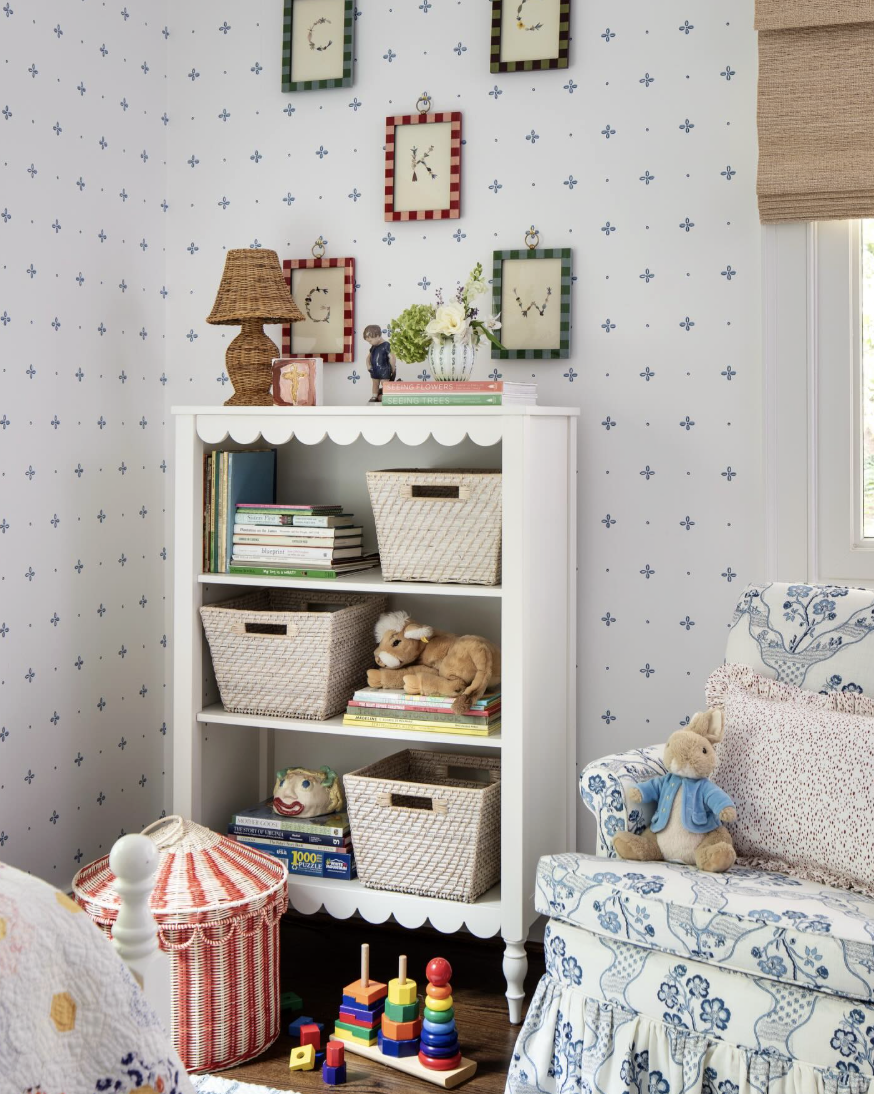 A children's room with a white bookcase, plush toys, and colorful books, decorated with framed stitched fabric art and a floral-patterned armchair.