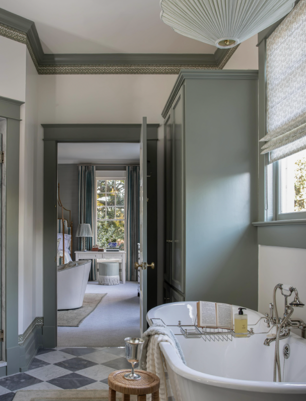 A bathroom with a clawfoot tub, a small wooden stool, and a host of natural light from a window, leading to a bedroom with a bed, curtains, and a view of trees outside.
